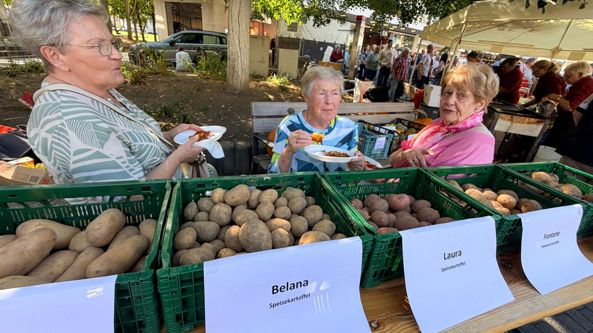 Drei Frauen sitzen hinter grünen Kisten, in denen Kartoffeln liegen.