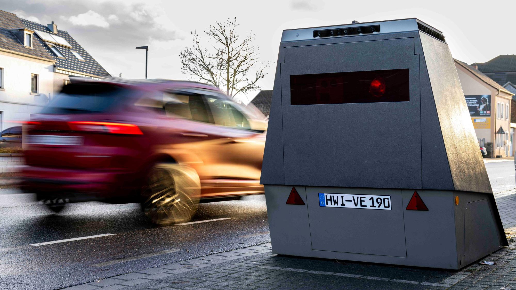 Ein rotes Auto fährt an einem Blitzeranhänger vorbei, der am Straßenrand abgestellt ist.