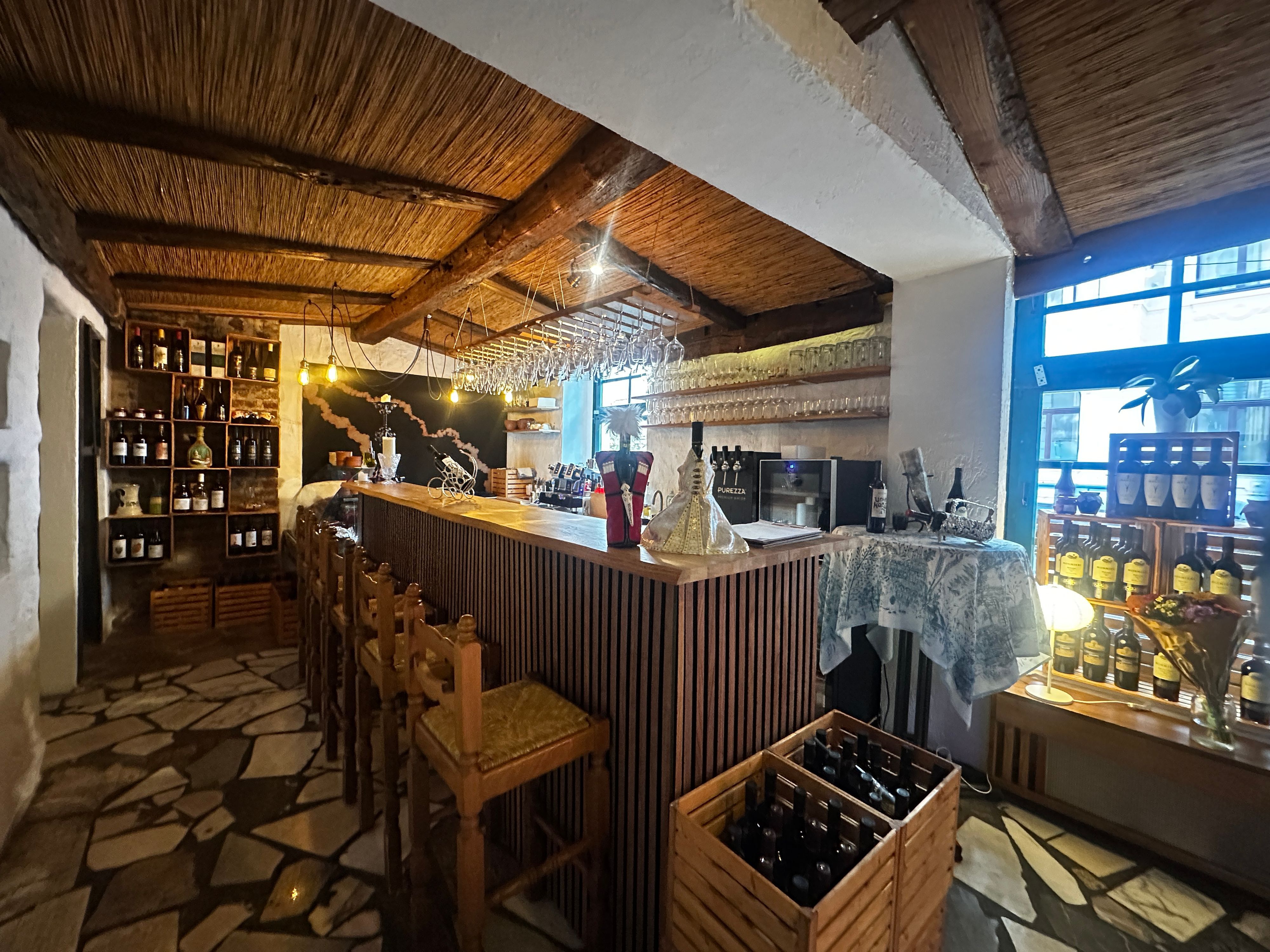 Innenansicht einer Weinbar mit Holzbalken, Theke, Barhockern und Regalen voller Flaschen und Gläser.