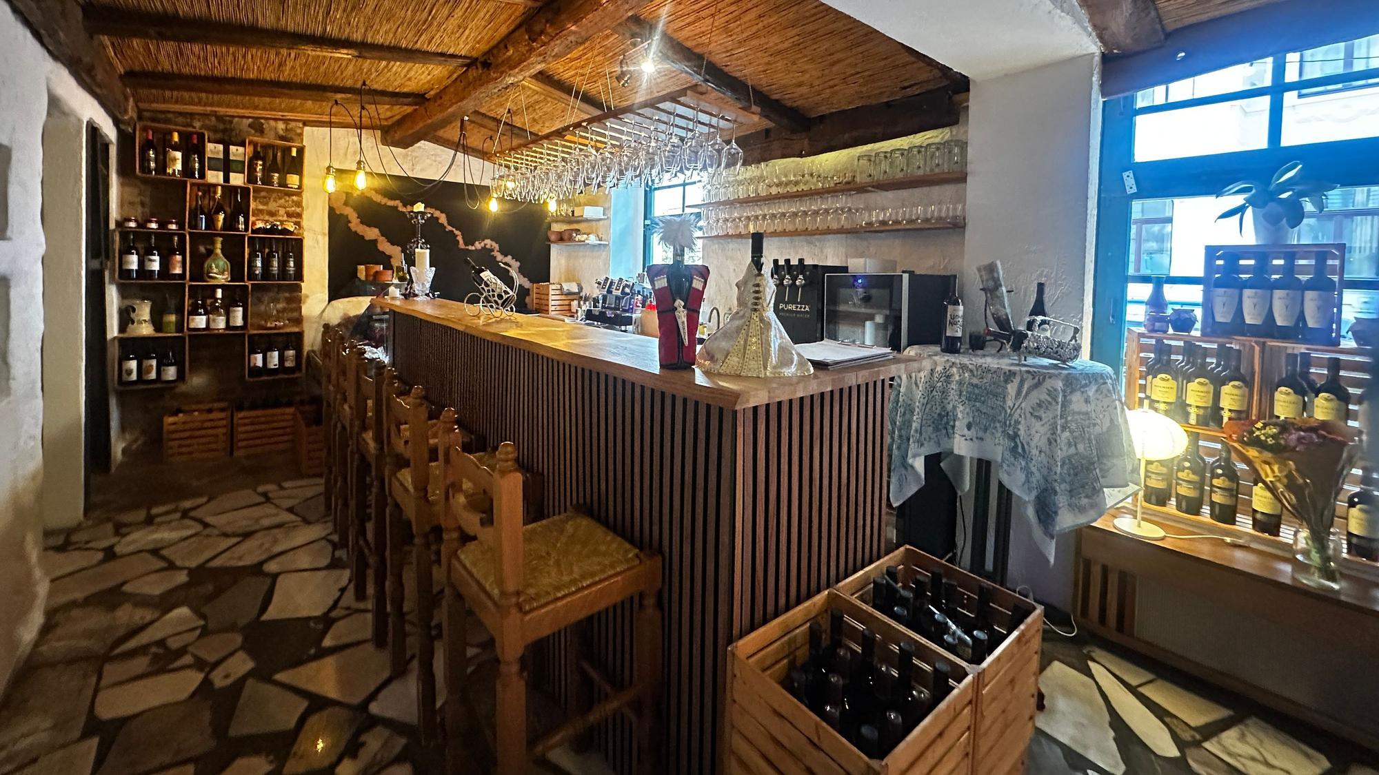 Innenansicht einer Weinbar mit Holzbalken, Theke, Barhockern und Regalen voller Flaschen und Gläser.
