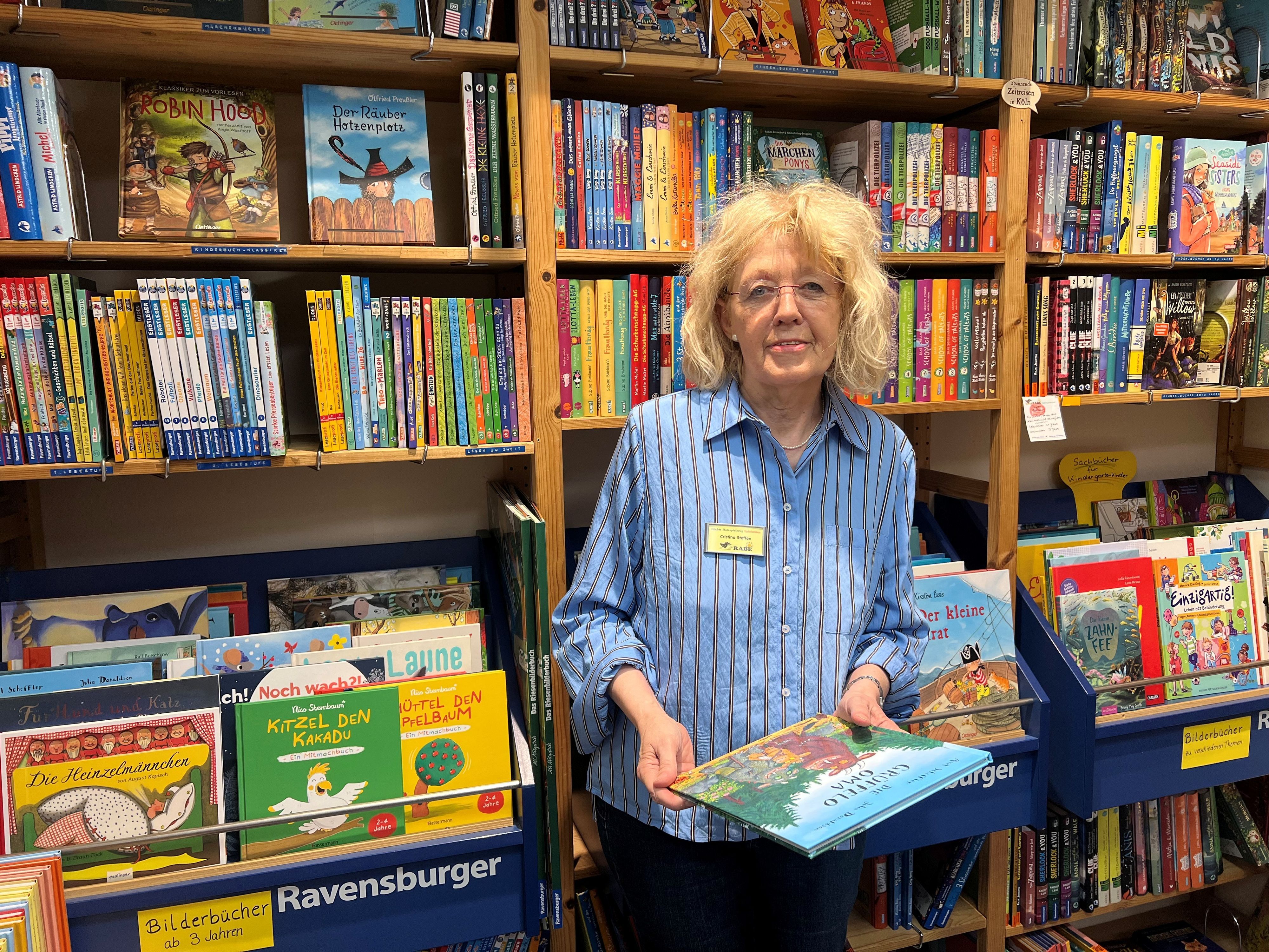Eine Frau steht zwischen Regalen voller Kinder- und Jugendbücher und hält ein Bilderbuch in den Händen.