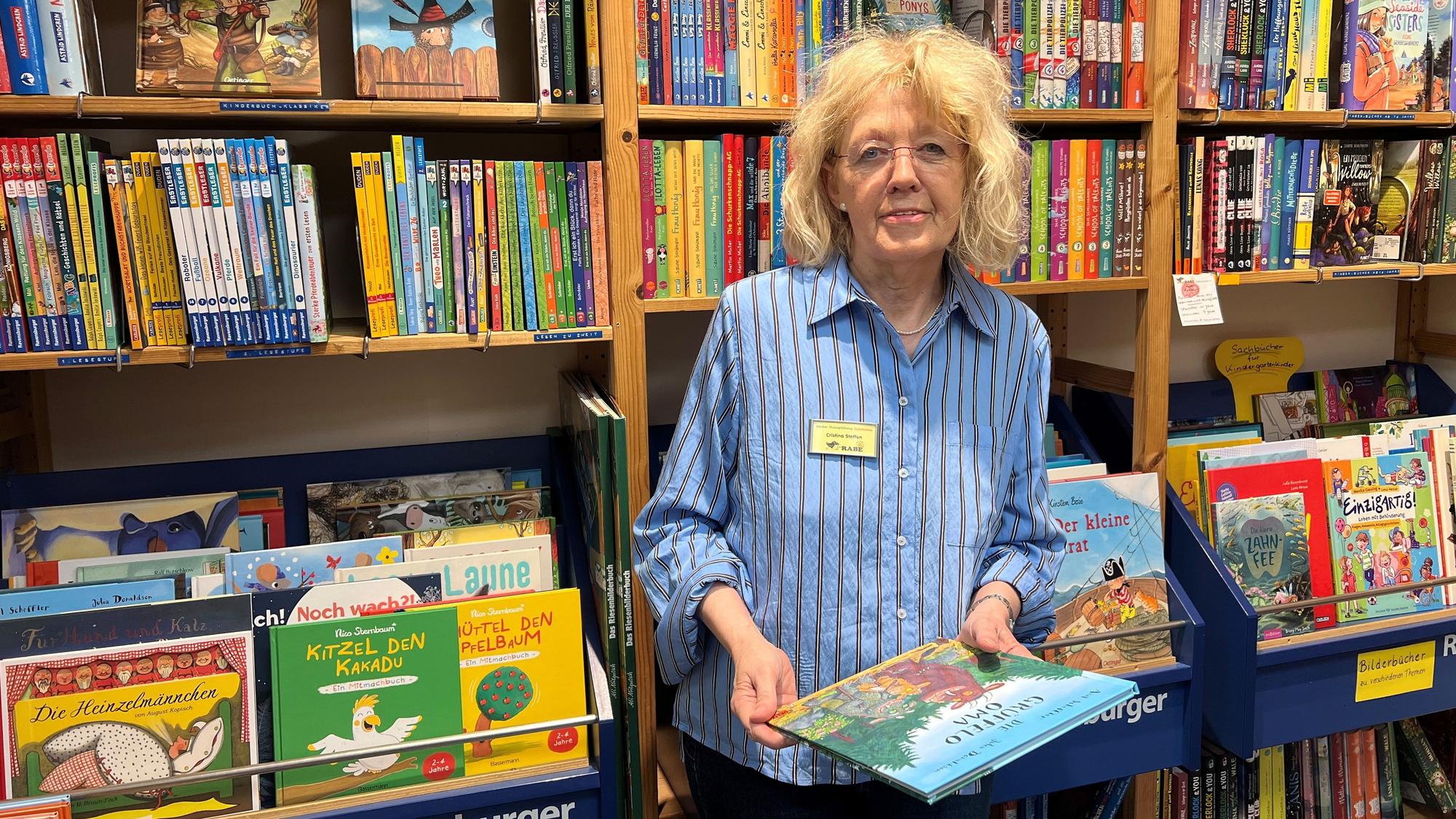 Eine Frau steht zwischen Regalen voller Kinder- und Jugendbücher und hält ein Bilderbuch in den Händen.