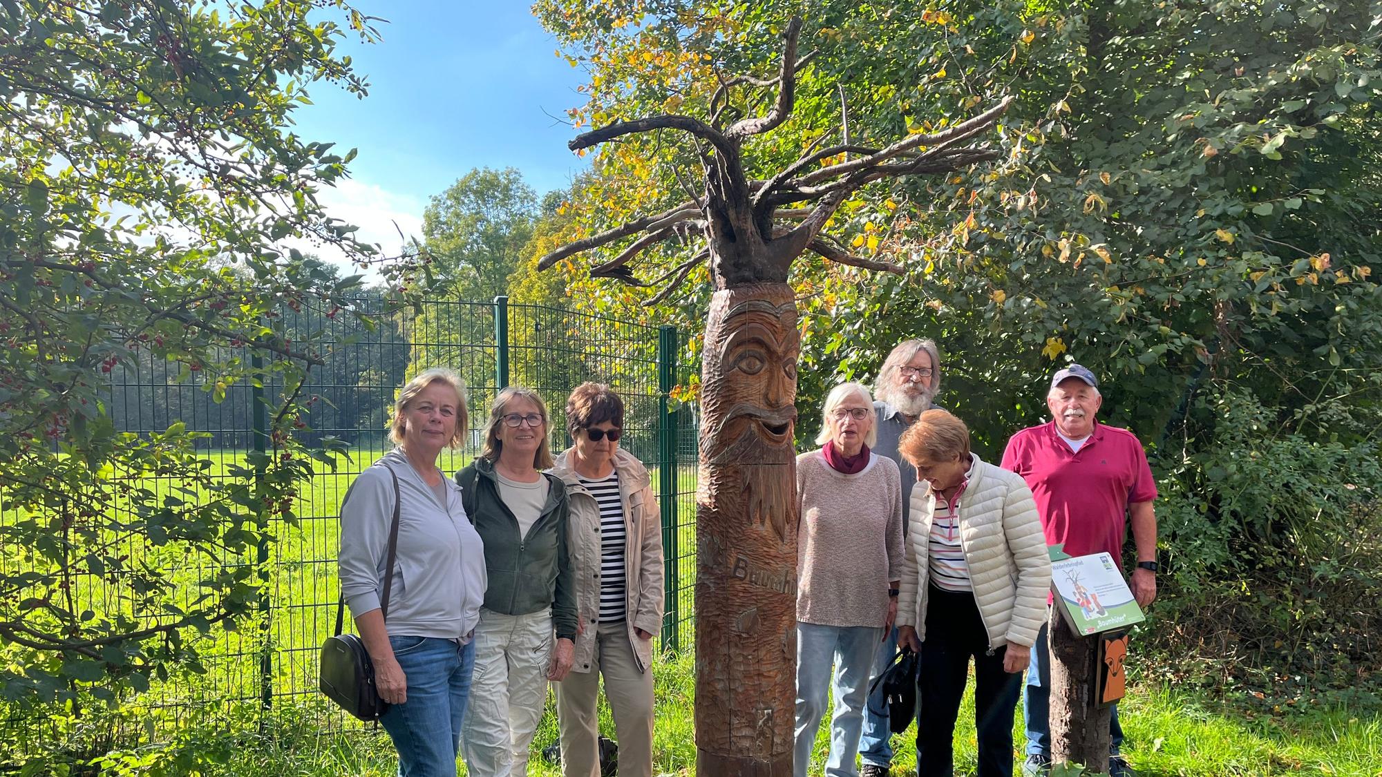Sieben Erwachsene stehen neben einer hohen, geschnitzten Holzfigur mit Gesicht und ausladenden Ästen. Im Hintergrund sind ein Zaun und ein grünes Gehege zu sehen.