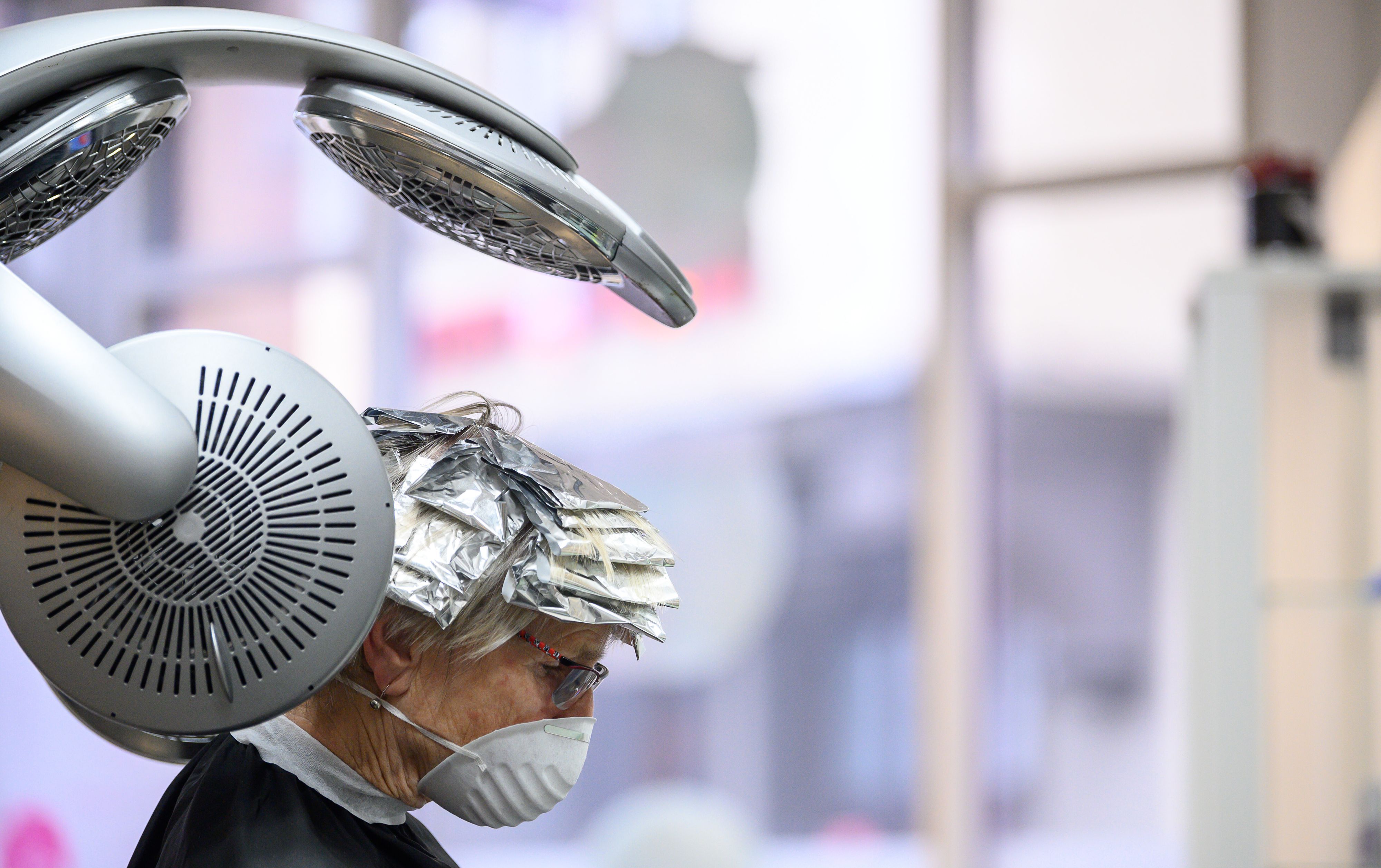 Eine Kundin mit Mund- und Nasenschutz sitzt in einem Friseursalon unter einem Trockengerät.