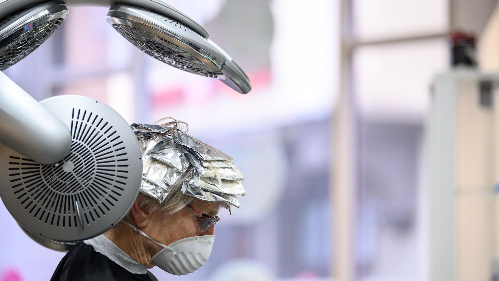 Eine Kundin mit Mund- und Nasenschutz sitzt in einem Friseursalon unter einem Trockengerät.