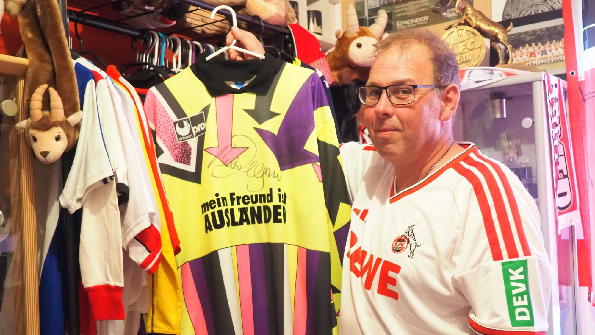 Rudi Faus steht vor einer Kleiderstange, an der unzählige Trikots hängen. Er hält eines hoch, auf dem "Mein Freund ist Ausländer" steht.