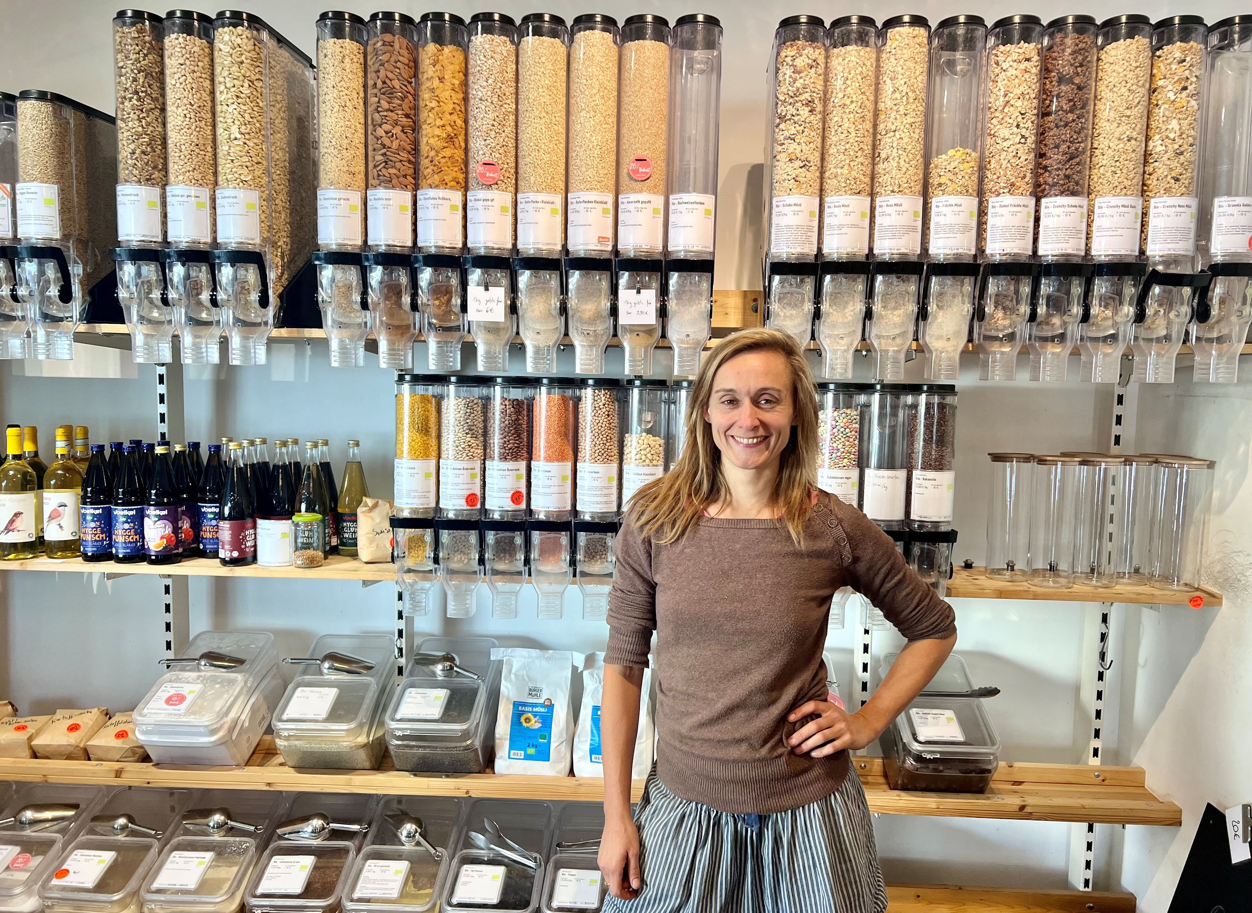 Eine Frau mit langem, mittelblondem Haar steht in einem Ladenlokal vor einem Regal, in dem gefüllte Unverpackt-Behälter zum Selbstabfüllen stehen.