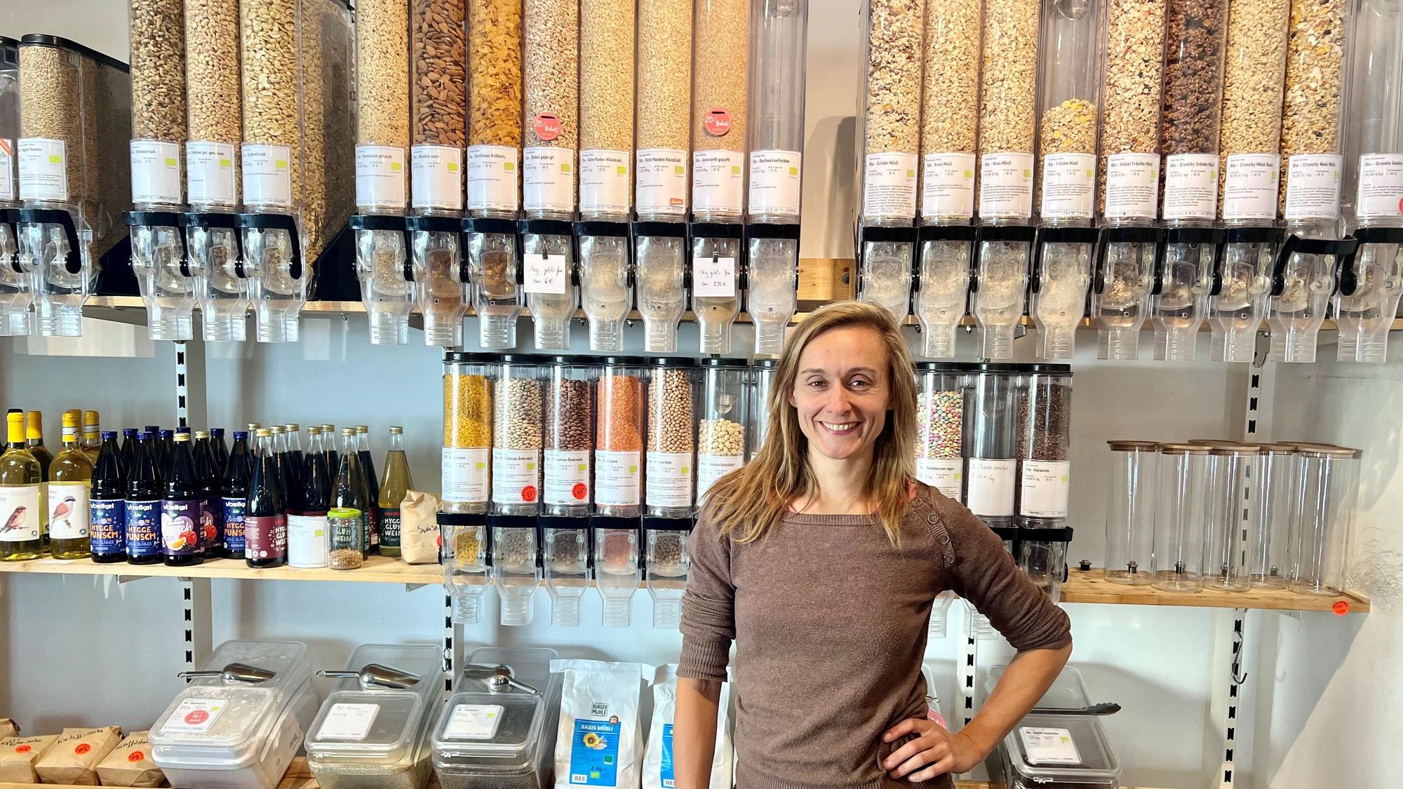 Eine Frau mit langem, mittelblondem Haar steht in einem Ladenlokal vor einem Regal, in dem gefüllte Unverpackt-Behälter zum Selbstabfüllen stehen.