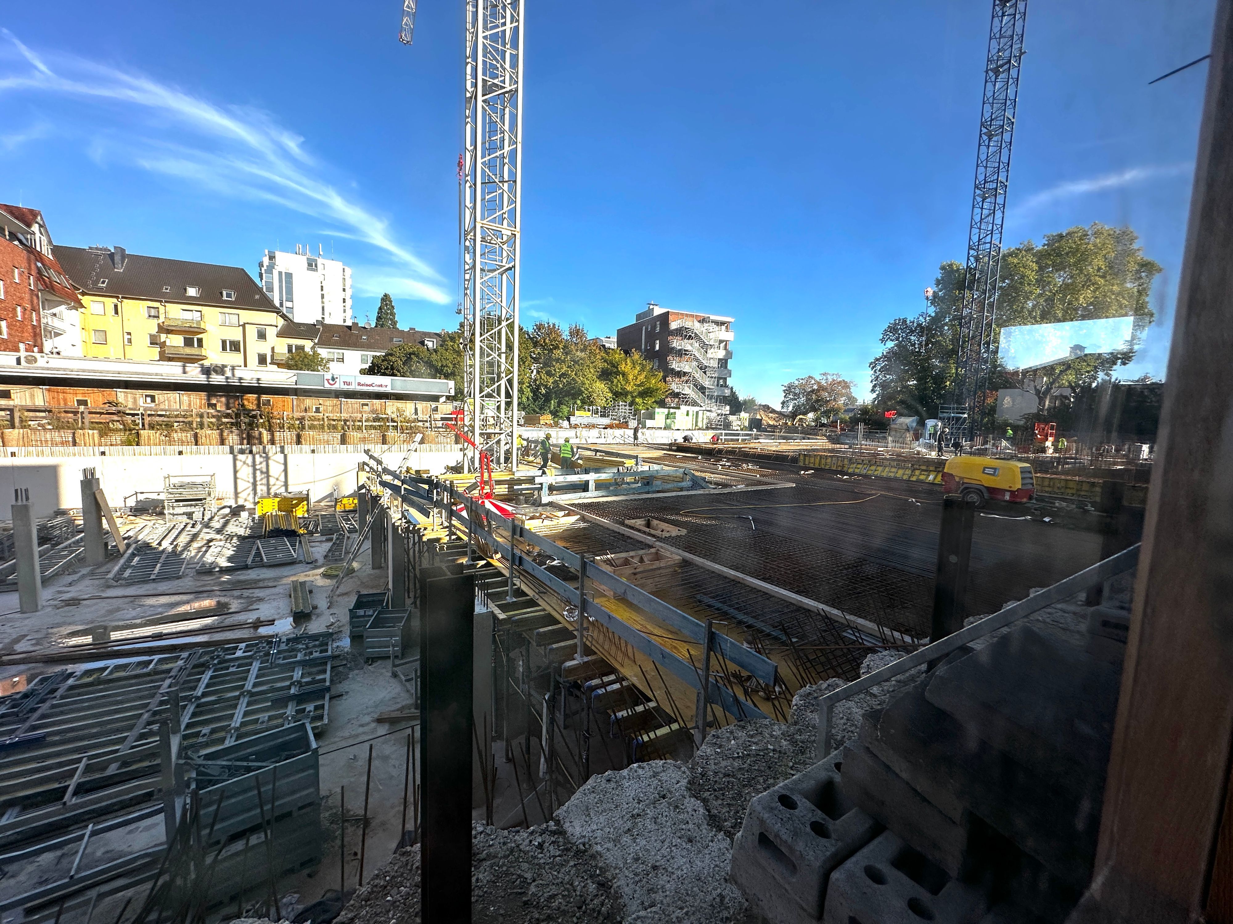 Zu sehen ist eine große Baustelle mit zwei Baukränen, einem teilweise betonierten Fundament und Stahlkonstruktionen, umgeben von Wohnhäusern und Bäumen.