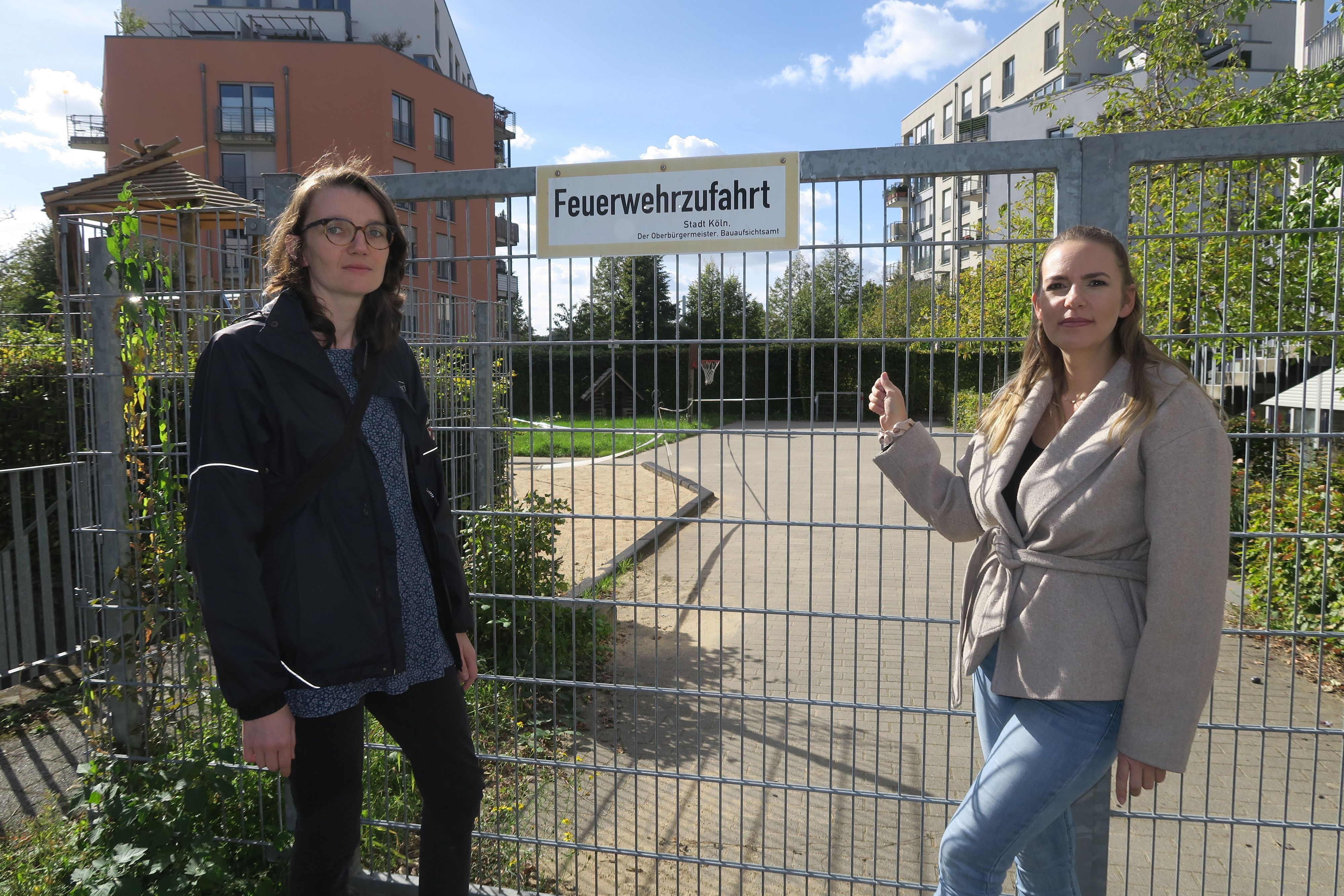 Zwei Frauen stehen vor einem abgesperrten Spielplatz; eine zeigt auf das Tor mit dem Schild „Feuerwehrzufahrt“.