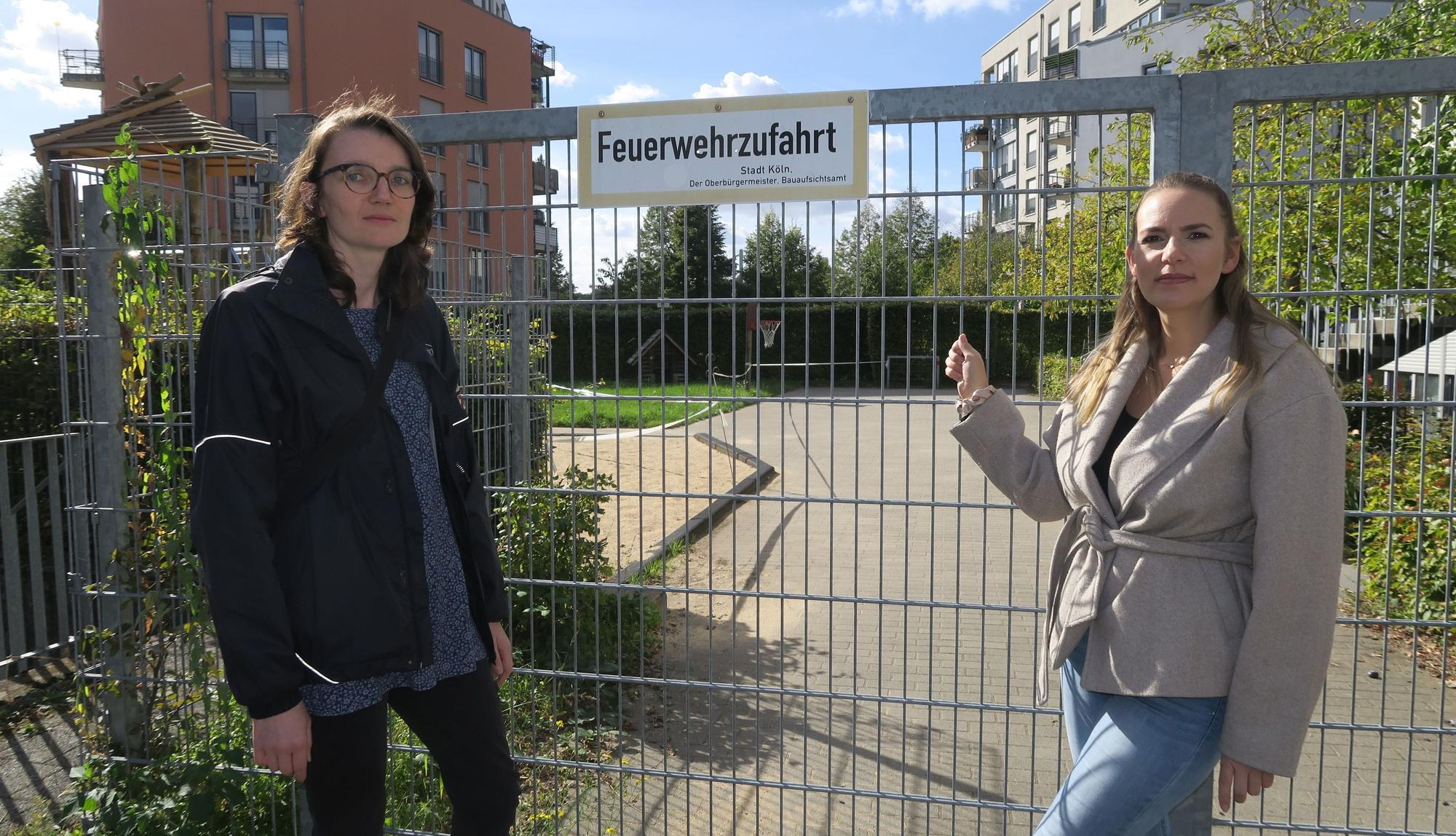Zwei Frauen stehen vor einem abgesperrten Spielplatz; eine zeigt auf das Tor mit dem Schild „Feuerwehrzufahrt“.