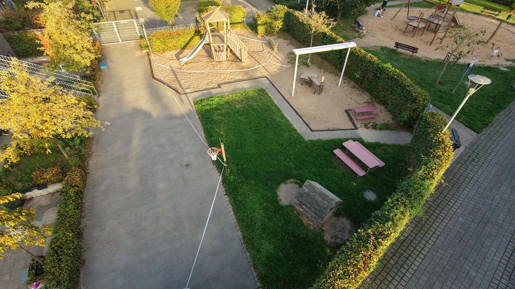 Ein Spielplatz mit Klettergerüst, Sandflächen und Sitzbänken ist aus der Vogelperspektive zu sehen; Absperrband verläuft um den Bereich.