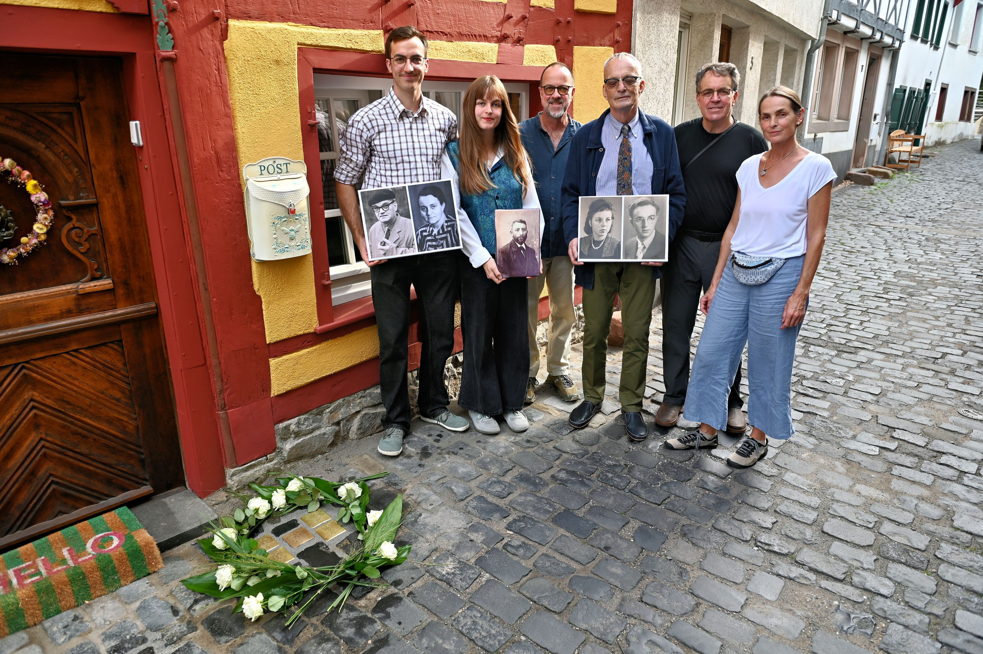 Sechs Personen stehen vor einem Fachwerkhaus, drei von ihnen halten Porträts ihrer Vorfahren in der Hand.