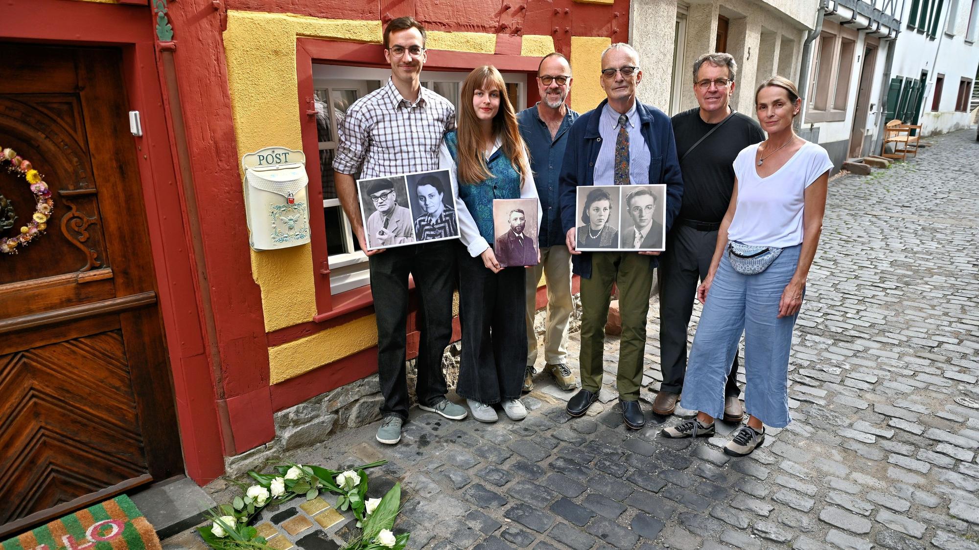 Sechs Personen stehen vor einem Fachwerkhaus, drei von ihnen halten Porträts ihrer Vorfahren in der Hand.