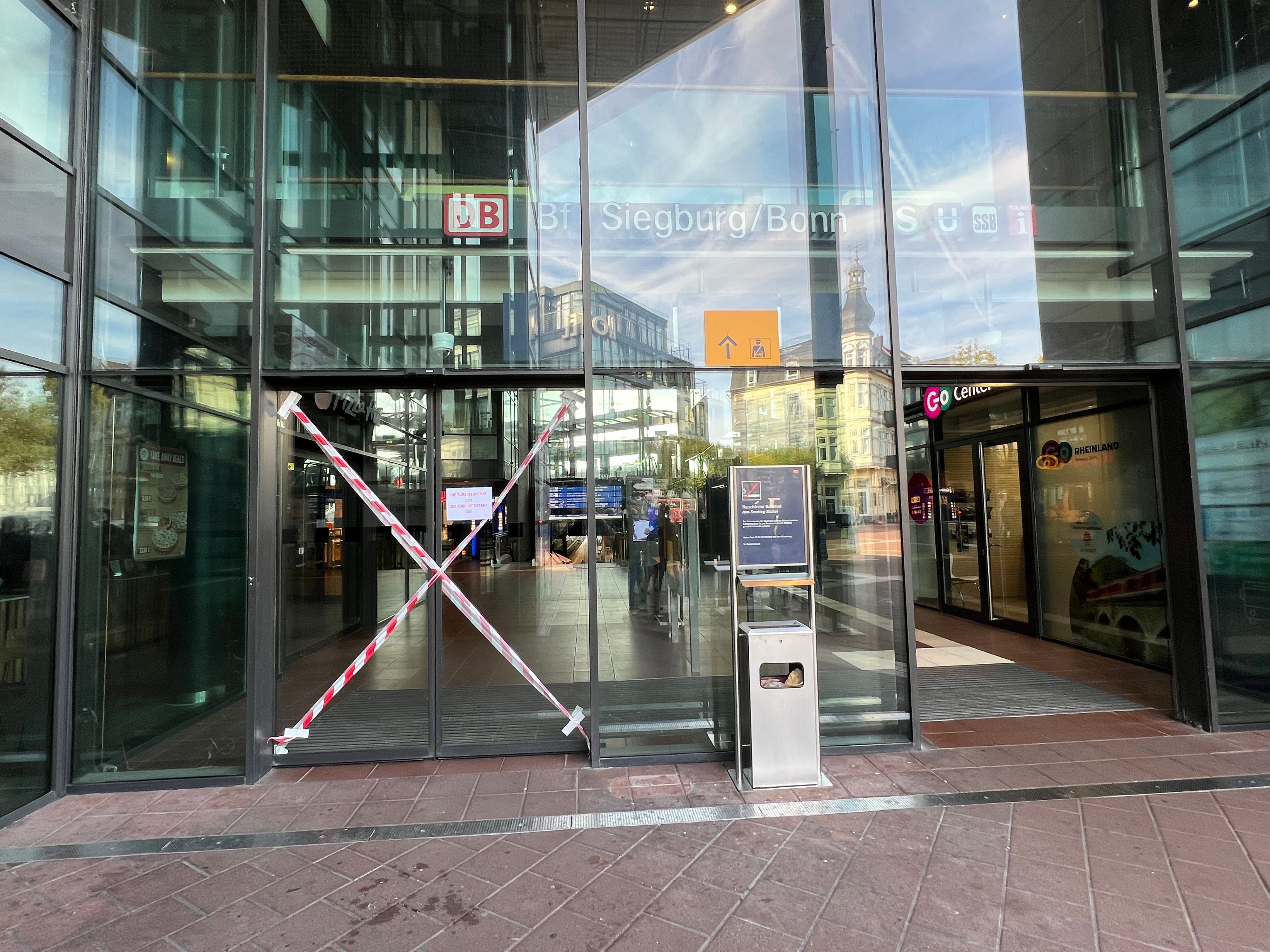 Mittlerweile professionell mit rot-weißem Flatterband als unpassierbar markiert ist eine der beiden elektrischen Schiebetüren zum Haupteingang des ICE-Bahnhofs in Siegburg. Sie ist seit mindestens drei Wochen (Stand 29.9.26) defekt.