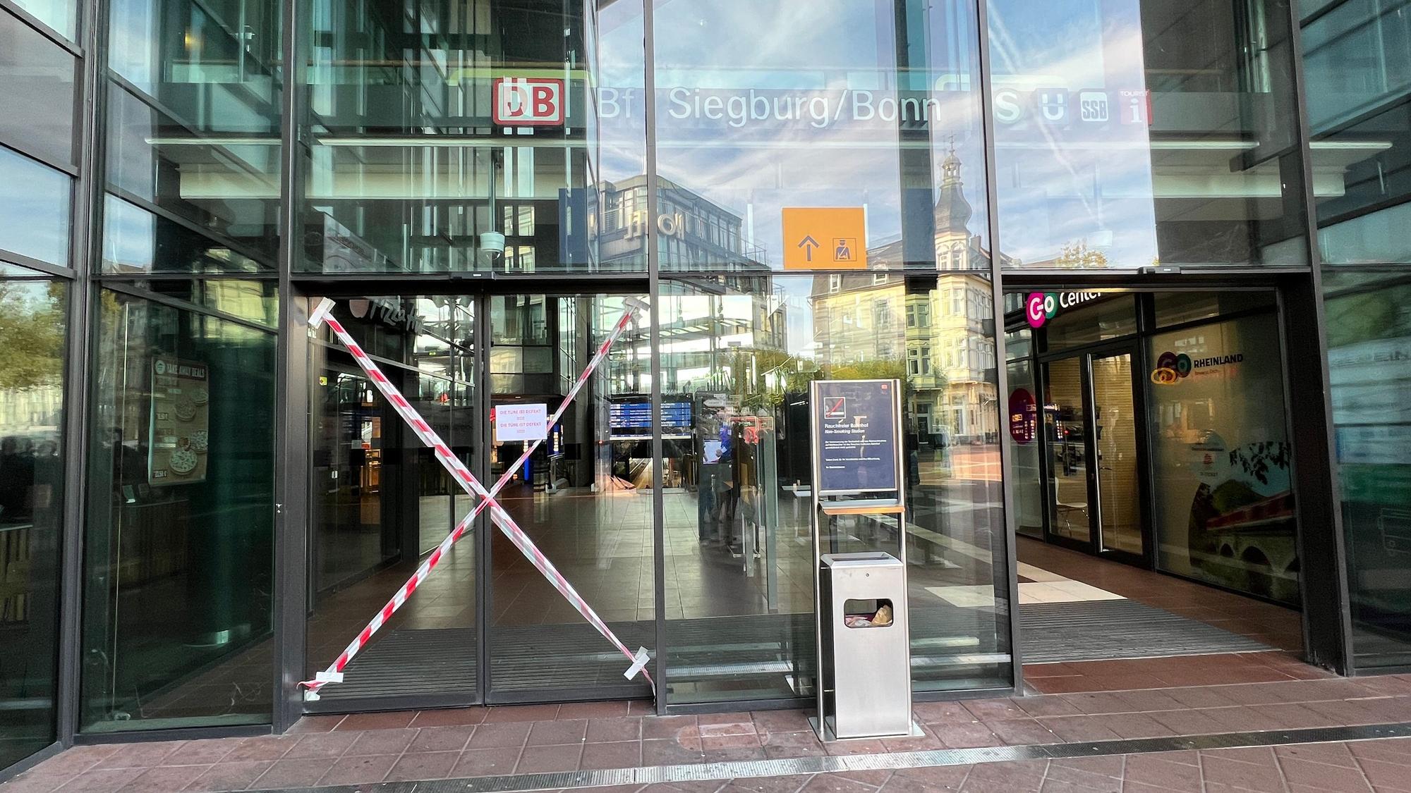 Mittlerweile professionell mit rot-weißem Flatterband als unpassierbar markiert ist eine der beiden elektrischen Schiebetüren zum Haupteingang des ICE-Bahnhofs in Siegburg. Sie ist seit mindestens drei Wochen (Stand 29.9.26) defekt.