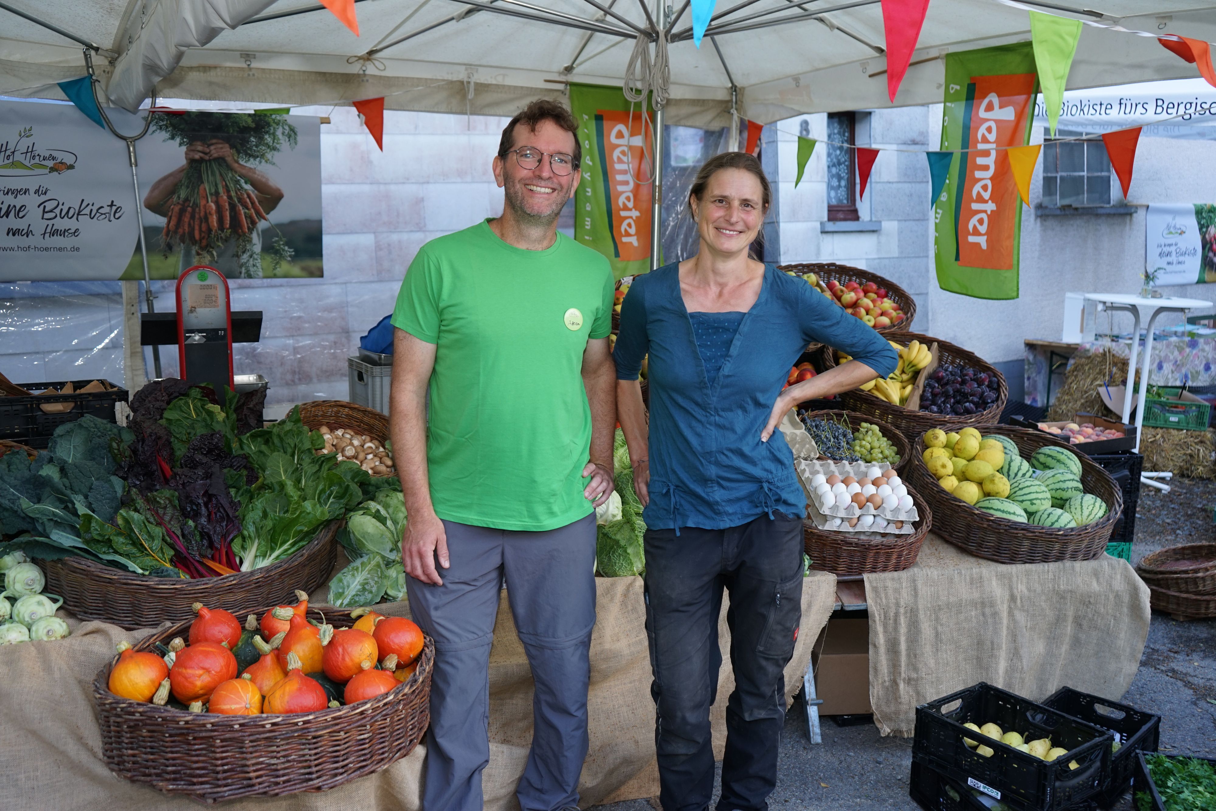 Eine Frau und ein Mann stehen an einem Stand umgeben von Körbern voller Obst und Gemüse.