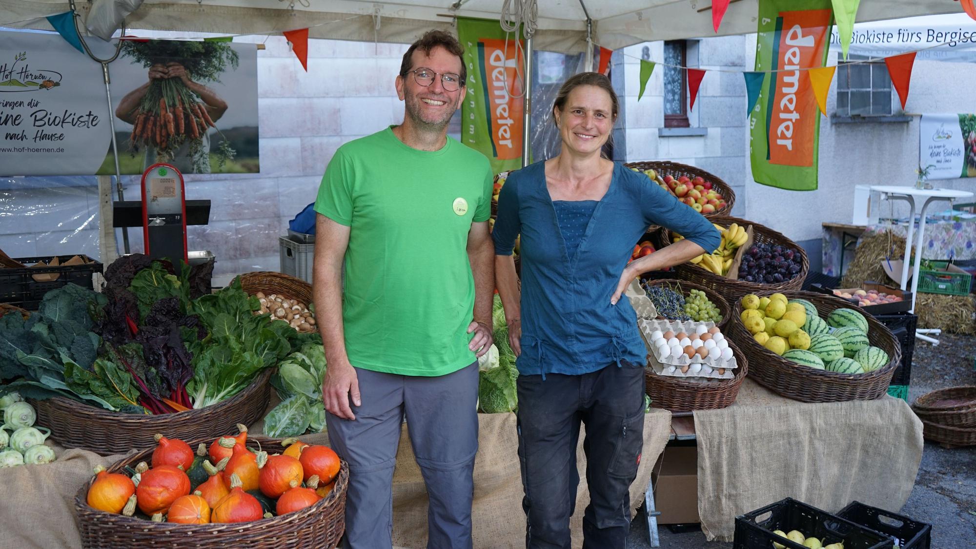 Eine Frau und ein Mann stehen an einem Stand umgeben von Körbern voller Obst und Gemüse.