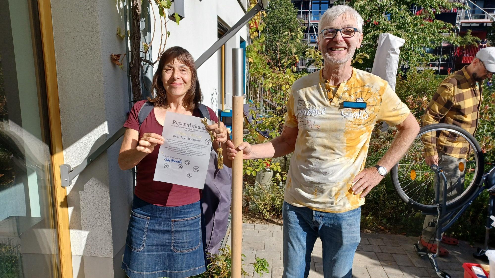 Eine Frau im Minirock steht neben einem Mann im gelben T-Shirt und Jeans. Er hält einen Holzstab in der Hand.