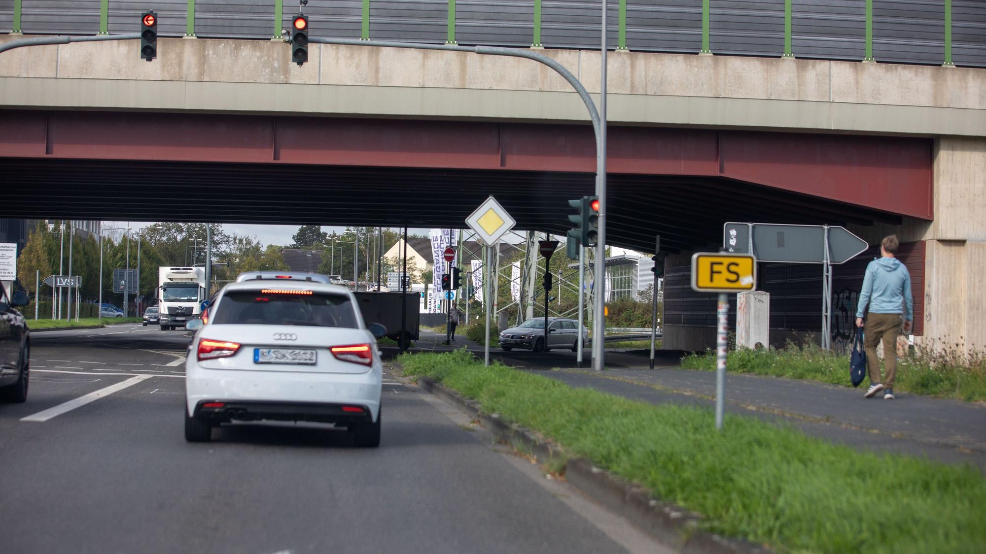 Eine stark befahrene Straßenkreuzung mit mehreren Fahrzeugen, Ampeln, Verkehrsschildern, Fahrbahnmarkierungen und einem Fußgänger neben den Straßen.