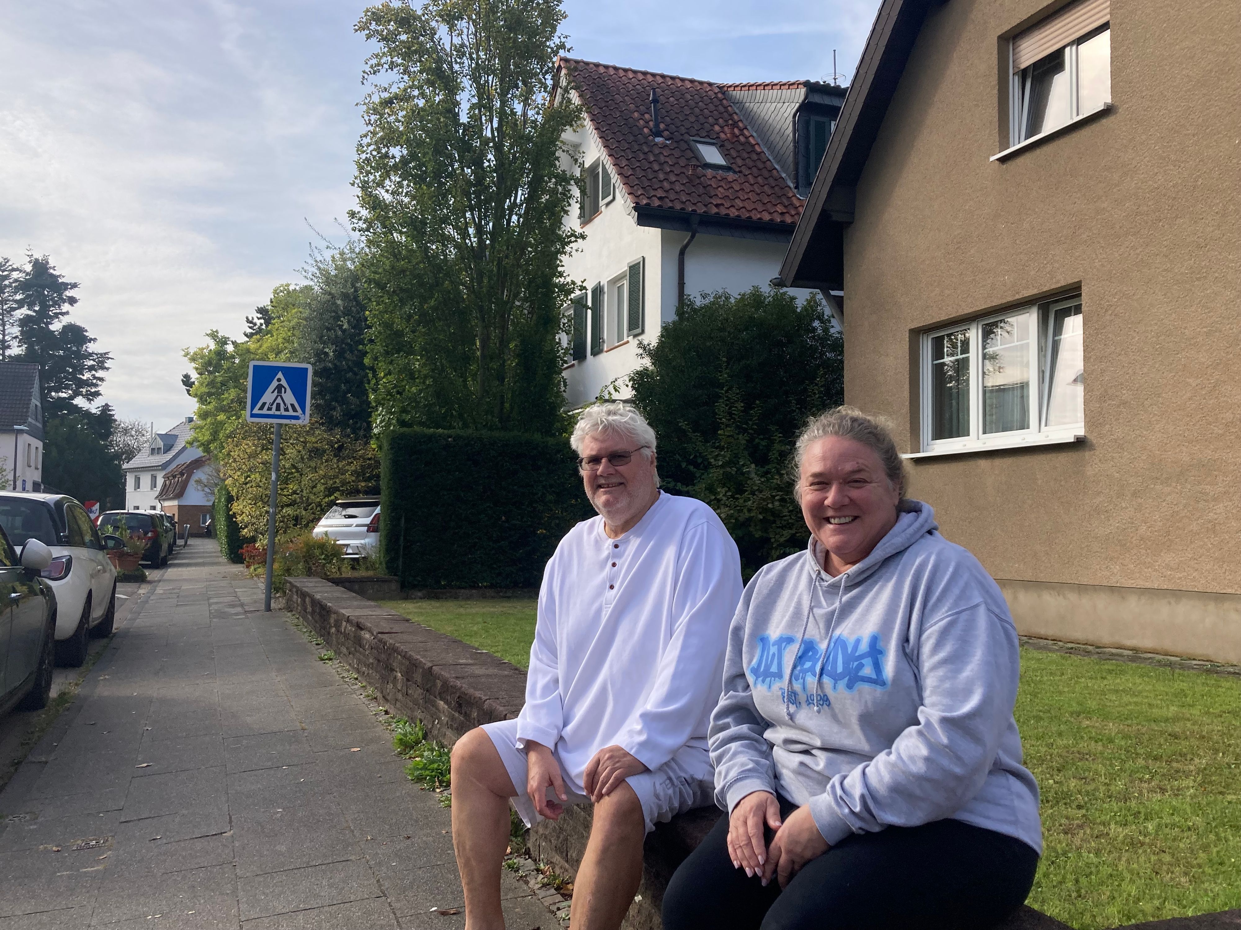 Zwei Menschen sitzen auf einer Mauer vor einem Vorgarten an einer Straße.