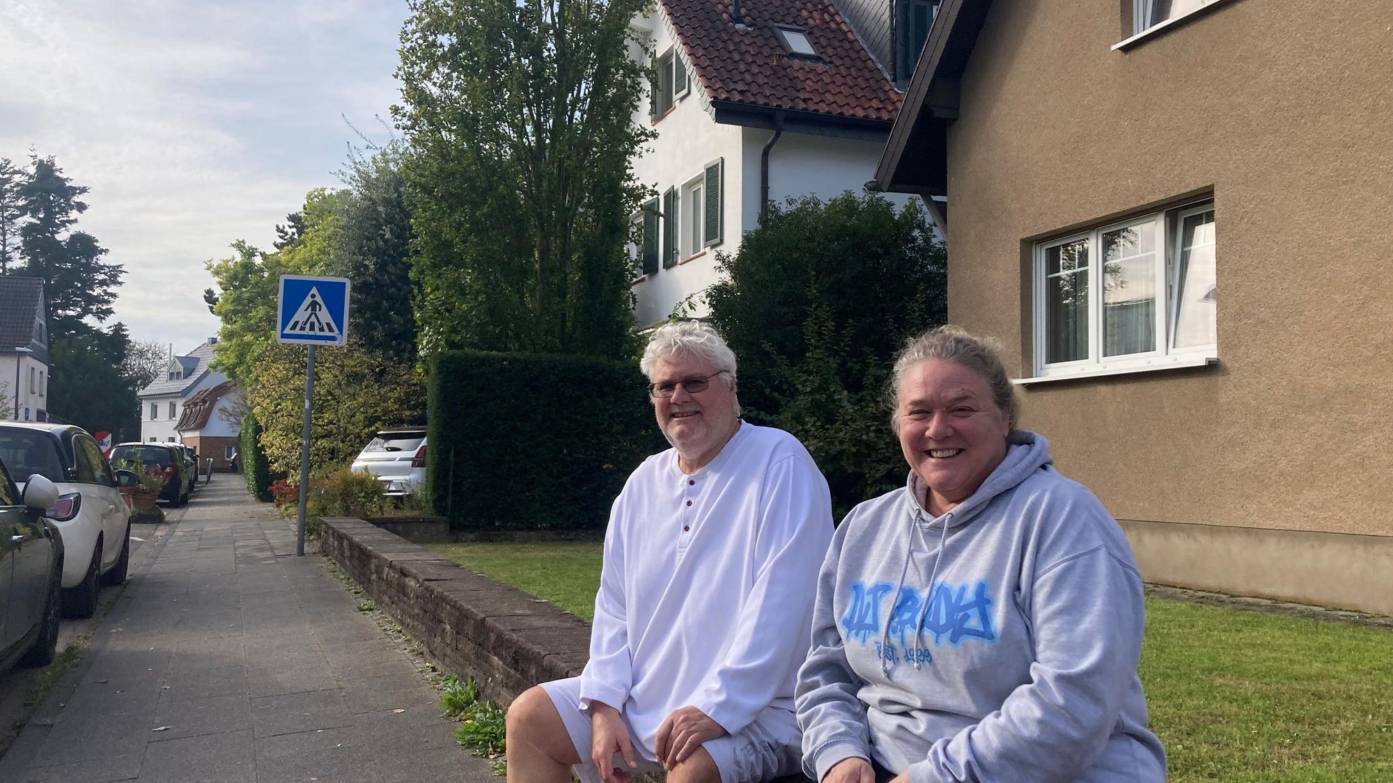 Zwei Menschen sitzen auf einer Mauer vor einem Vorgarten an einer Straße.