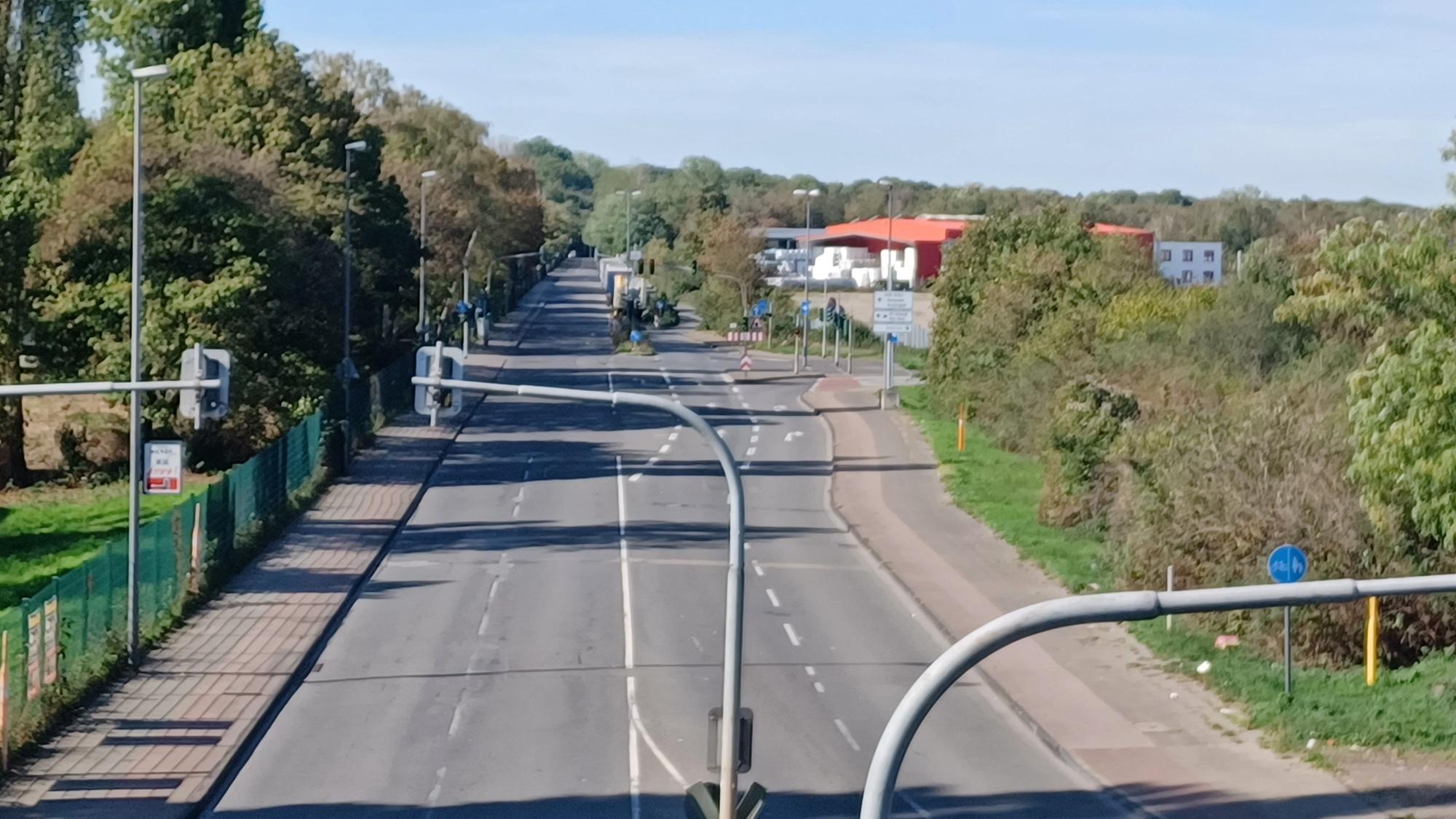 Das Foto zeigt eine mehrspurige Straße mit Gehwegen, Ampeln und Verkehrsschildern, die von Bäumen und Gebäuden gesäumt ist.