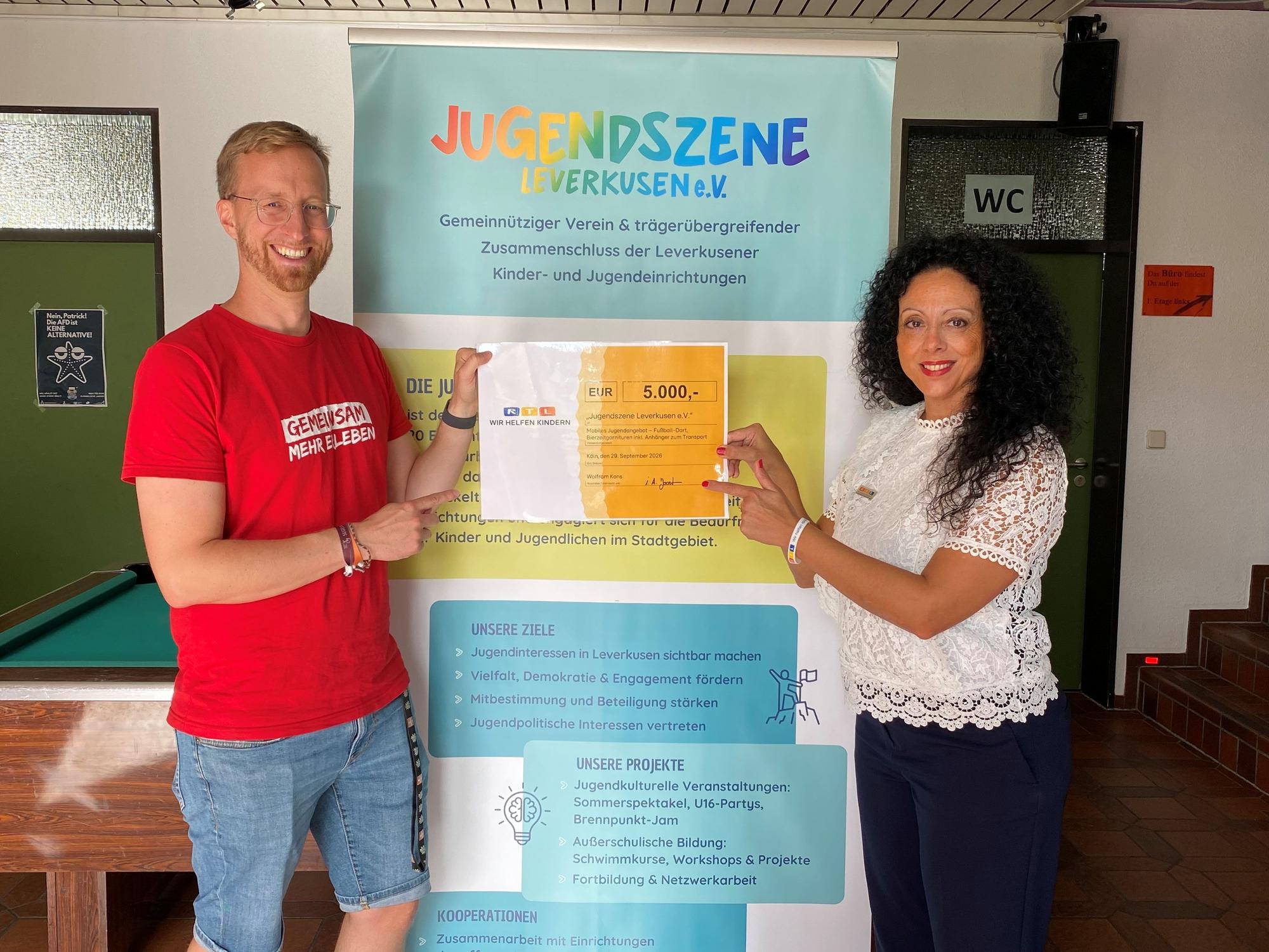 Das Bild zeigt zwei Menschen mit einem Check von 5000 Euro für die „Jugendszene Leverkusen“.