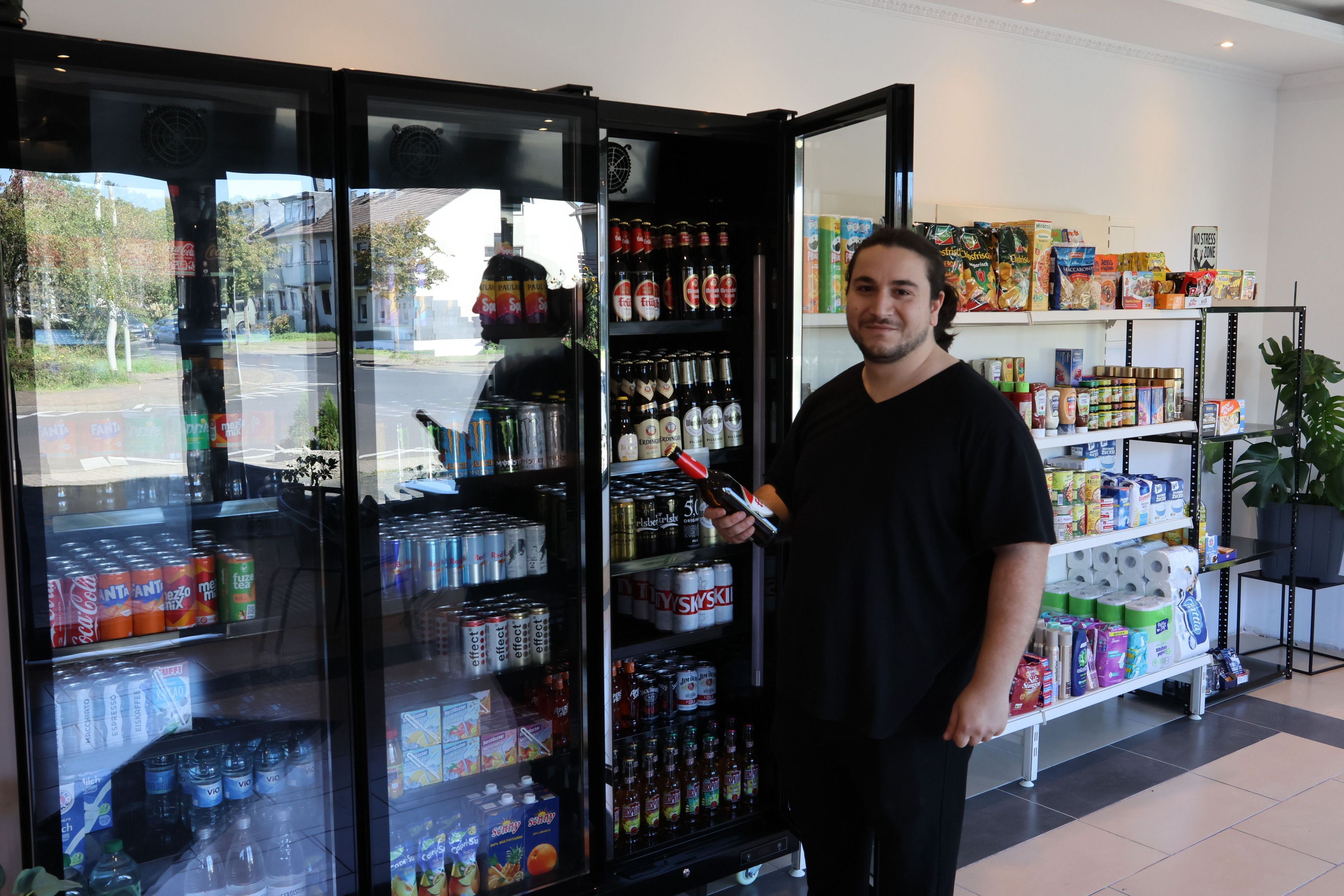 Zu sehen ist Safak Mutlu, erhält vor einem geöffneten Kühlschrank eine Flasche in der Hand.