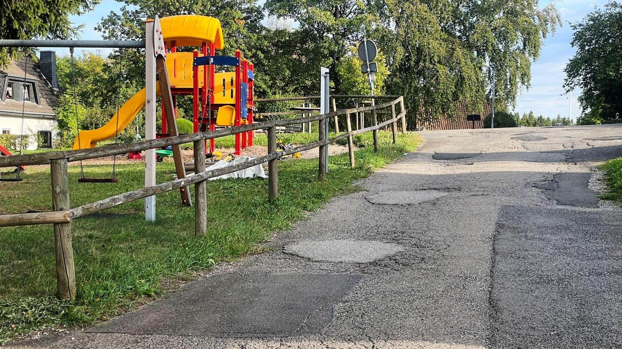 An einem Spielplatz führt eine mit etlichen Flickstellen versehene Asphalstraße vorbei.