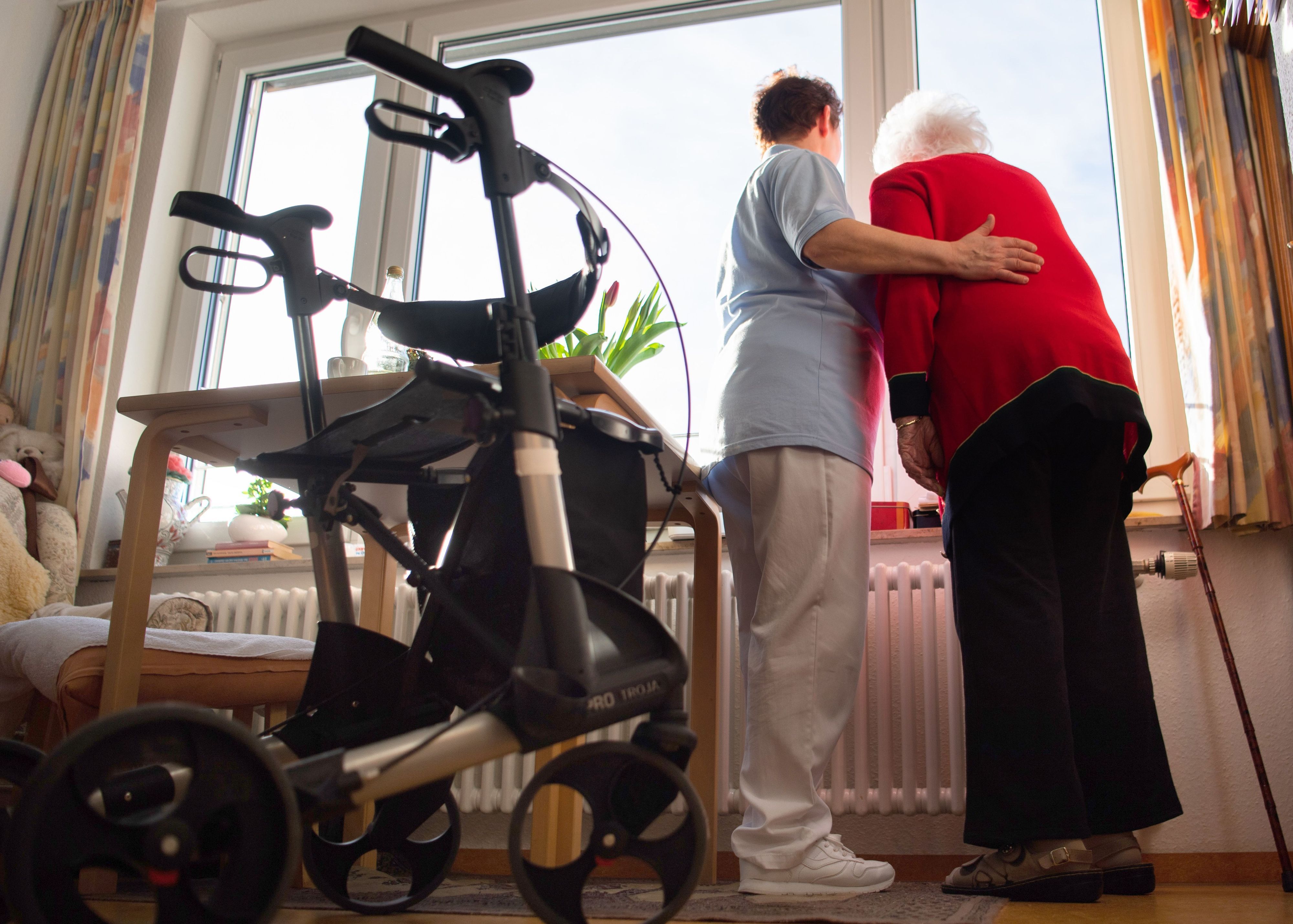 Eine Pflegerin und eine Heimbewohnerin schauen zusammen aus einem Fenster im Wohnbereich des Pflegeheims. Im Vordergrund steht ein Rollator. (Symbolbild)