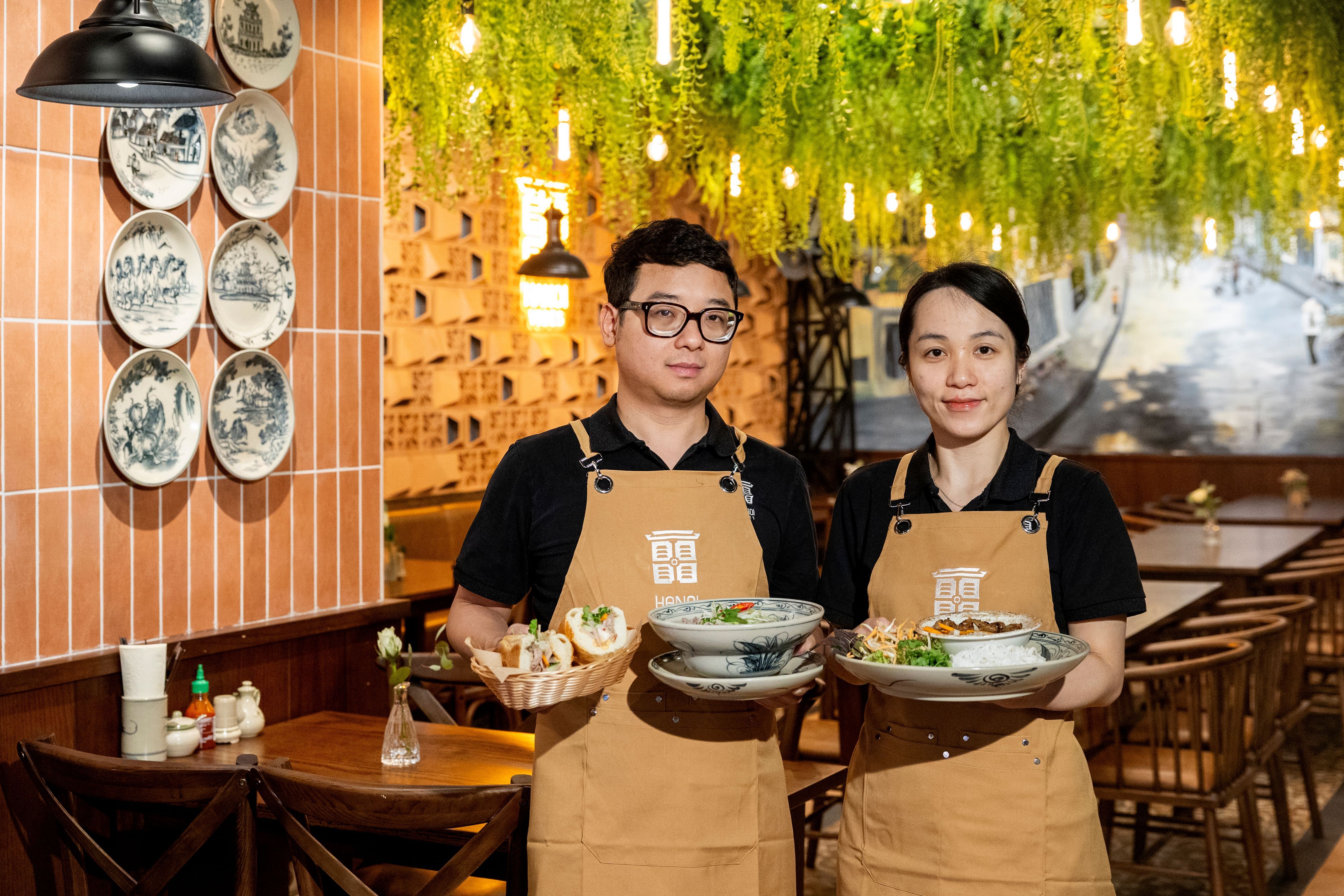 Ngoc Bich Tran und Viet Long Nguyen stehen in ihrem Restaurant und halten angerichtete Teller in der Hand. Sie tragen beige Schürzen.