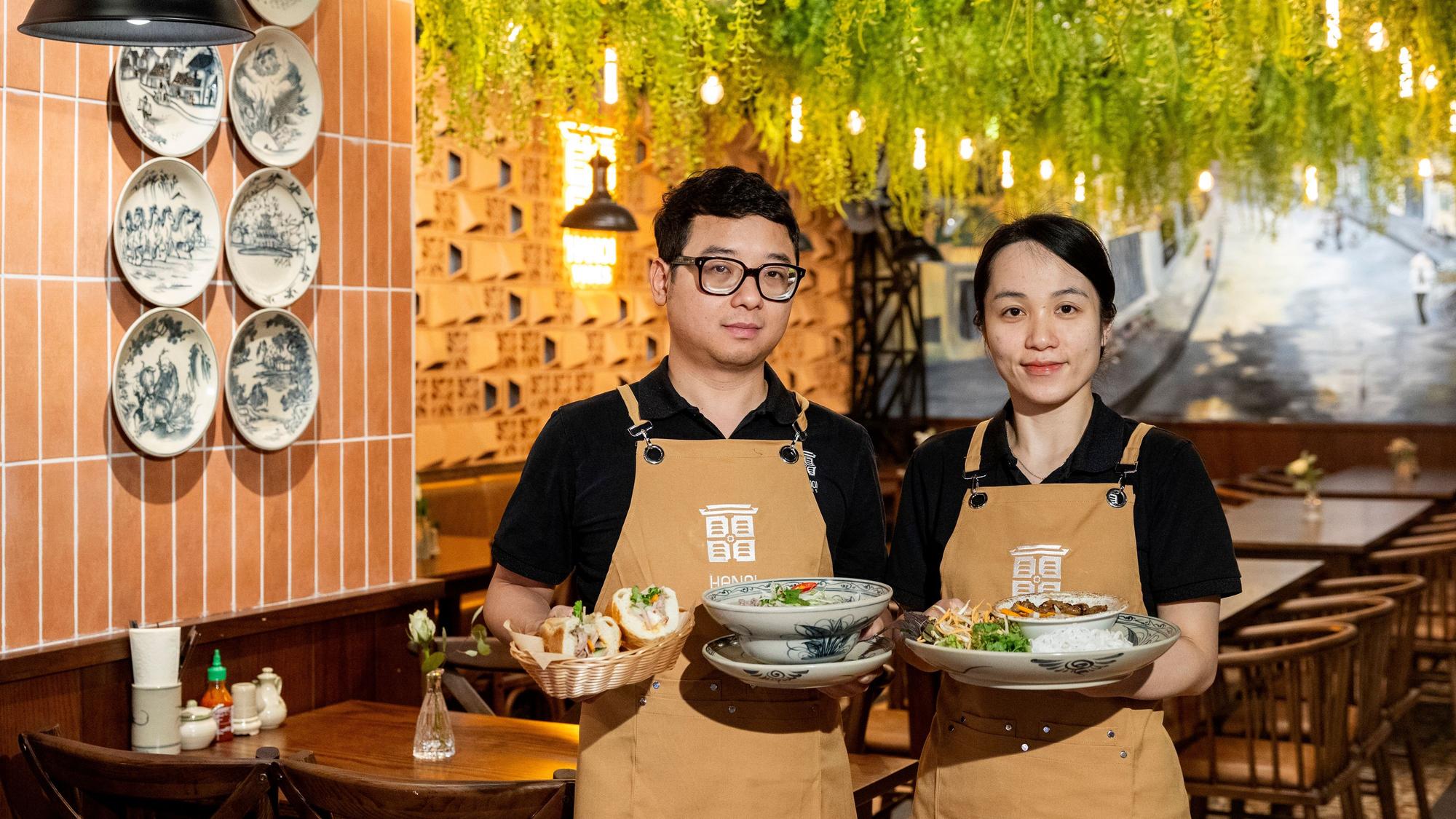 Ngoc Bich Tran und Viet Long Nguyen stehen in ihrem Restaurant und halten angerichtete Teller in der Hand. Sie tragen beige Schürzen.