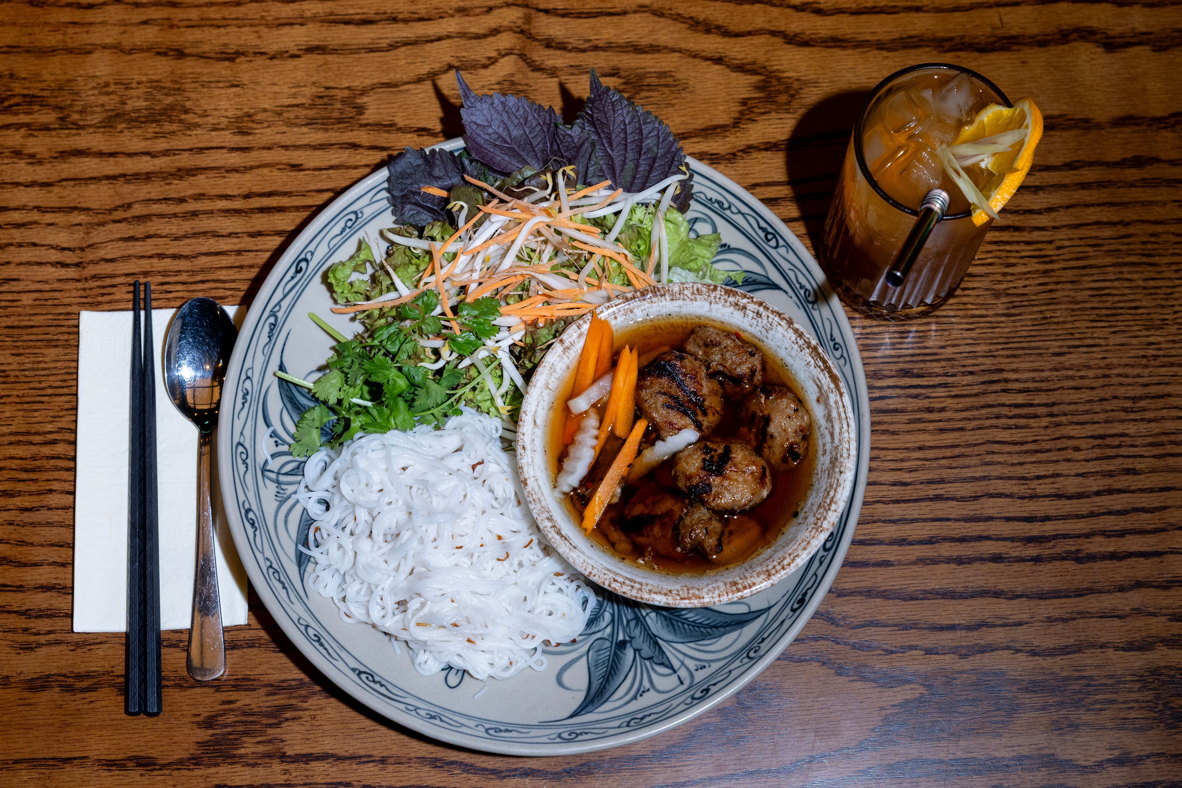 Bun Cha Hanoi: gegrilltes Schweinefleisch mit würzigen Hackfleischbällchen, Reisnudeln, Salat und Kräutern, angerichtet auf einem Teller, danben steht eine Limonade