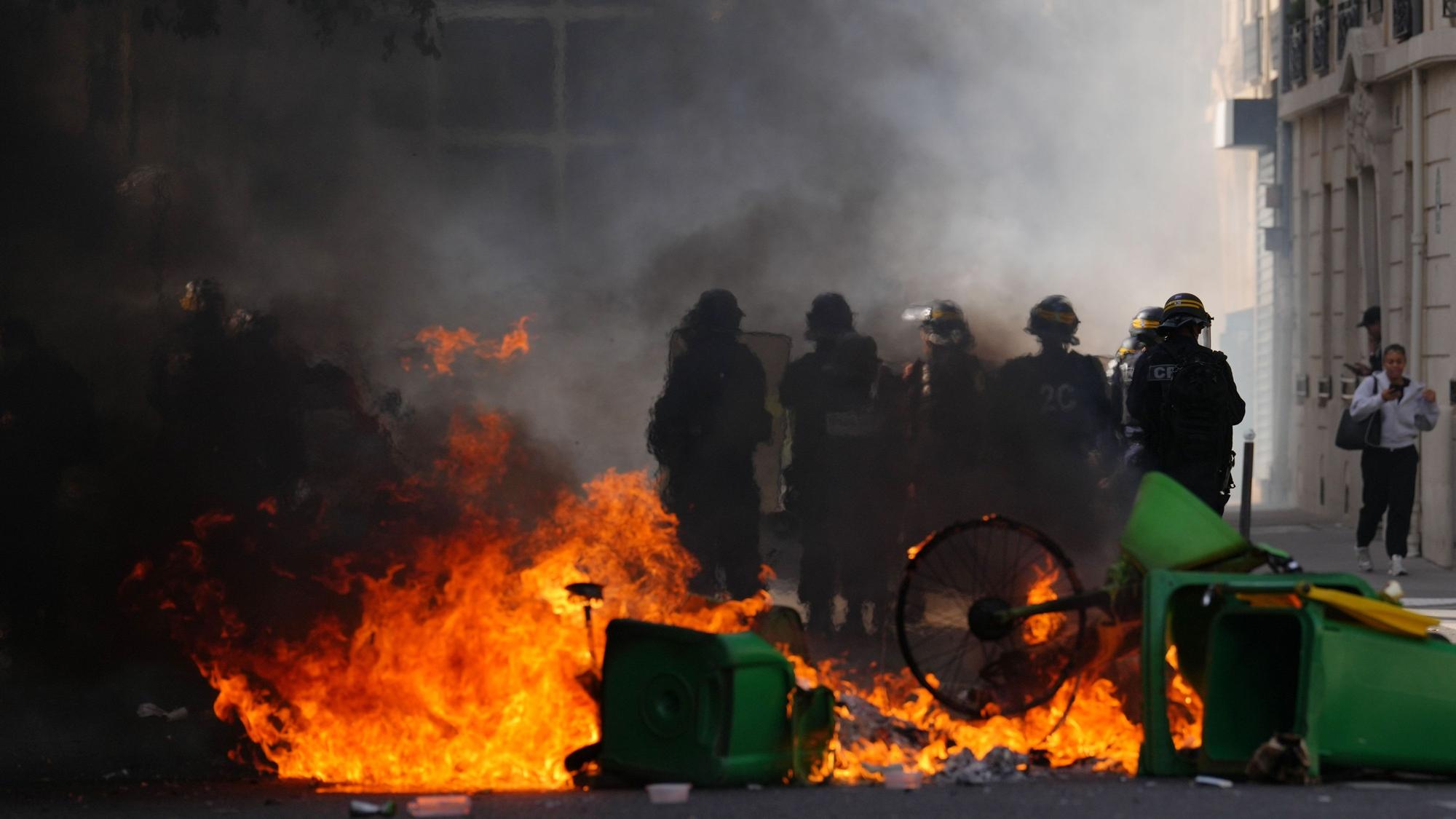 Polizisten stehen vor brennendem Müll, während sie bei einer Demonstration gegen unterfinanzierte Schulen Schülerinnen und Schülern gegenüberstehen.