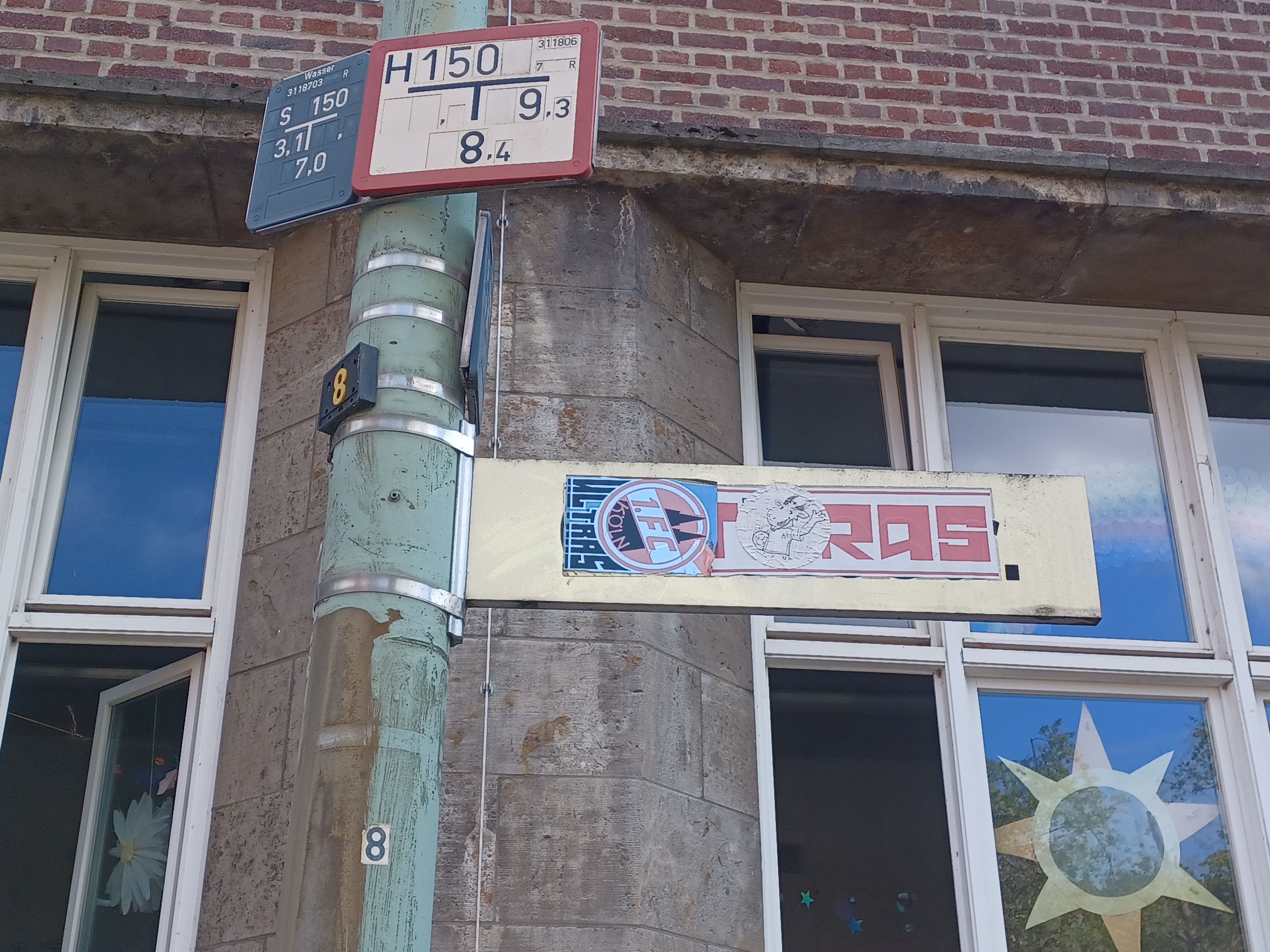 Ein mit FC‑Ultra-Aufklebern verunstaltetes Straßenschild steht vor einem Schulgebäude.
