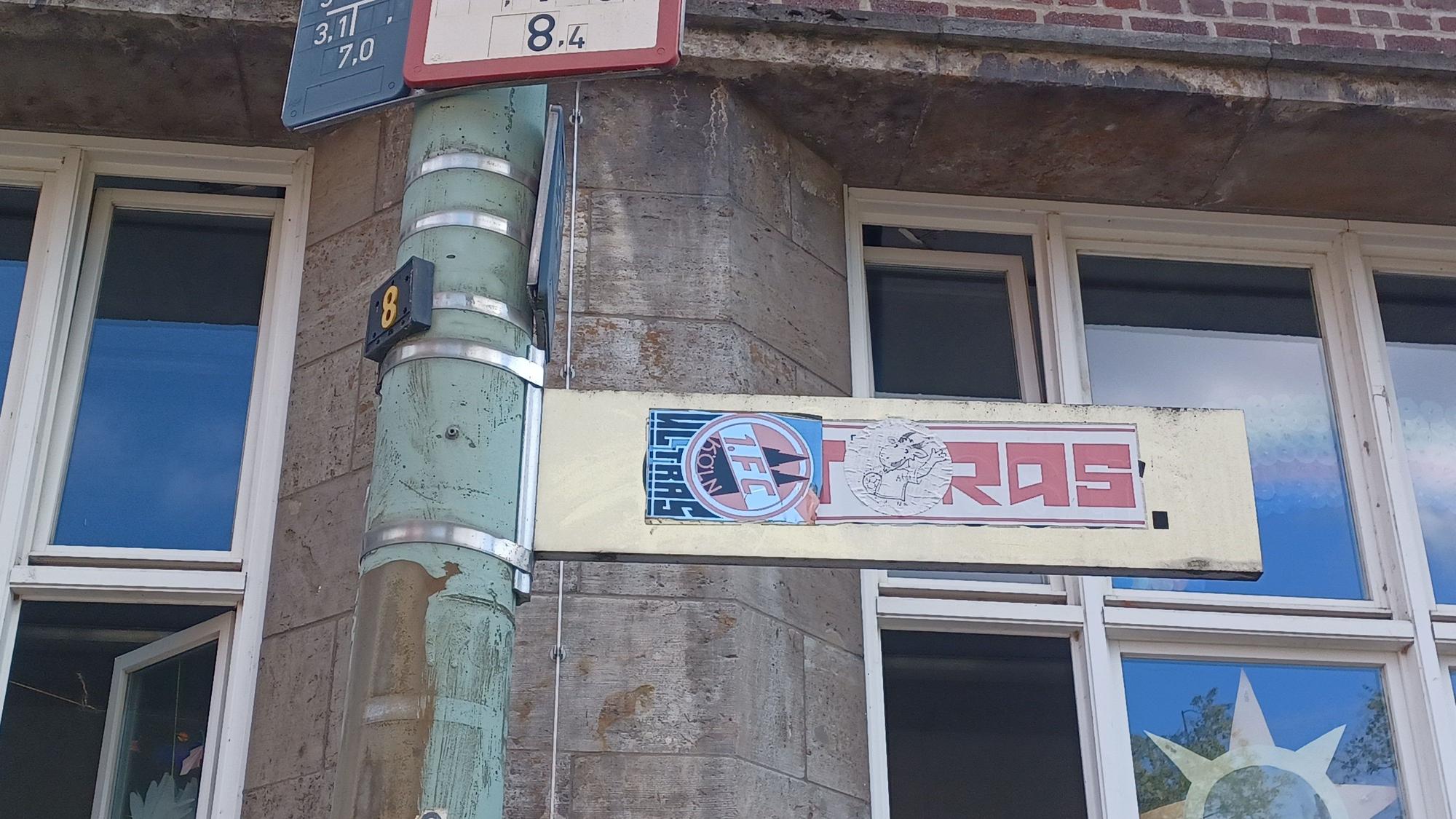 Ein mit FC‑Ultra-Aufklebern verunstaltetes Straßenschild steht vor einem Schulgebäude.