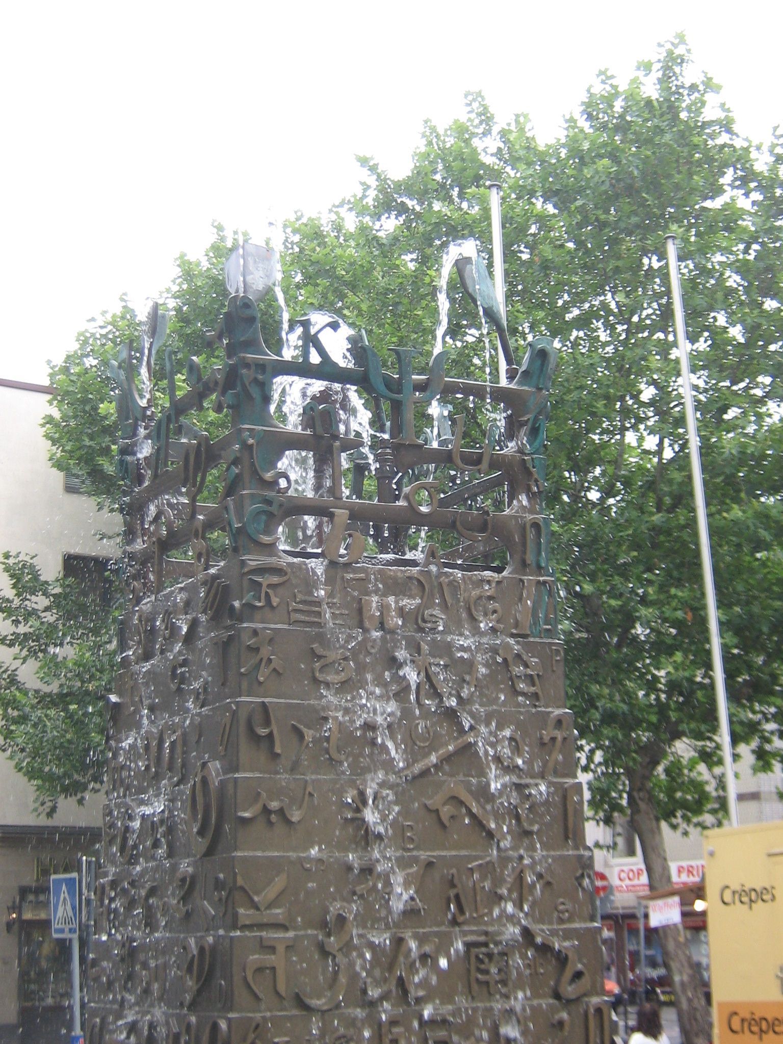 Ein sich konisch verjüngender Metallbrunnen speit Wasser. Er ist mit vielerlei Schriftzeichen verziert.