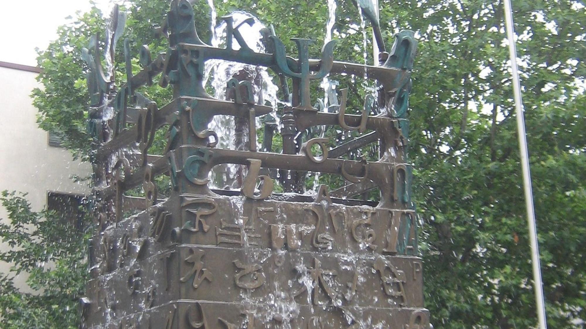 Ein sich konisch verjüngender Metallbrunnen speit Wasser. Er ist mit vielerlei Schriftzeichen verziert.