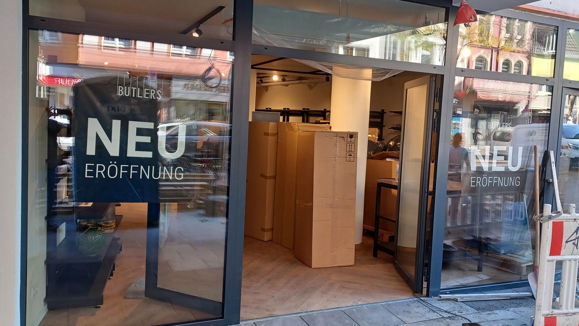 Ein Ladenlokal mit geöffnetem Eingang. An den Fenstern steht „NEU ERÖFFNUNG“; im Innenraum sind Kartons und Möbel zu sehen.