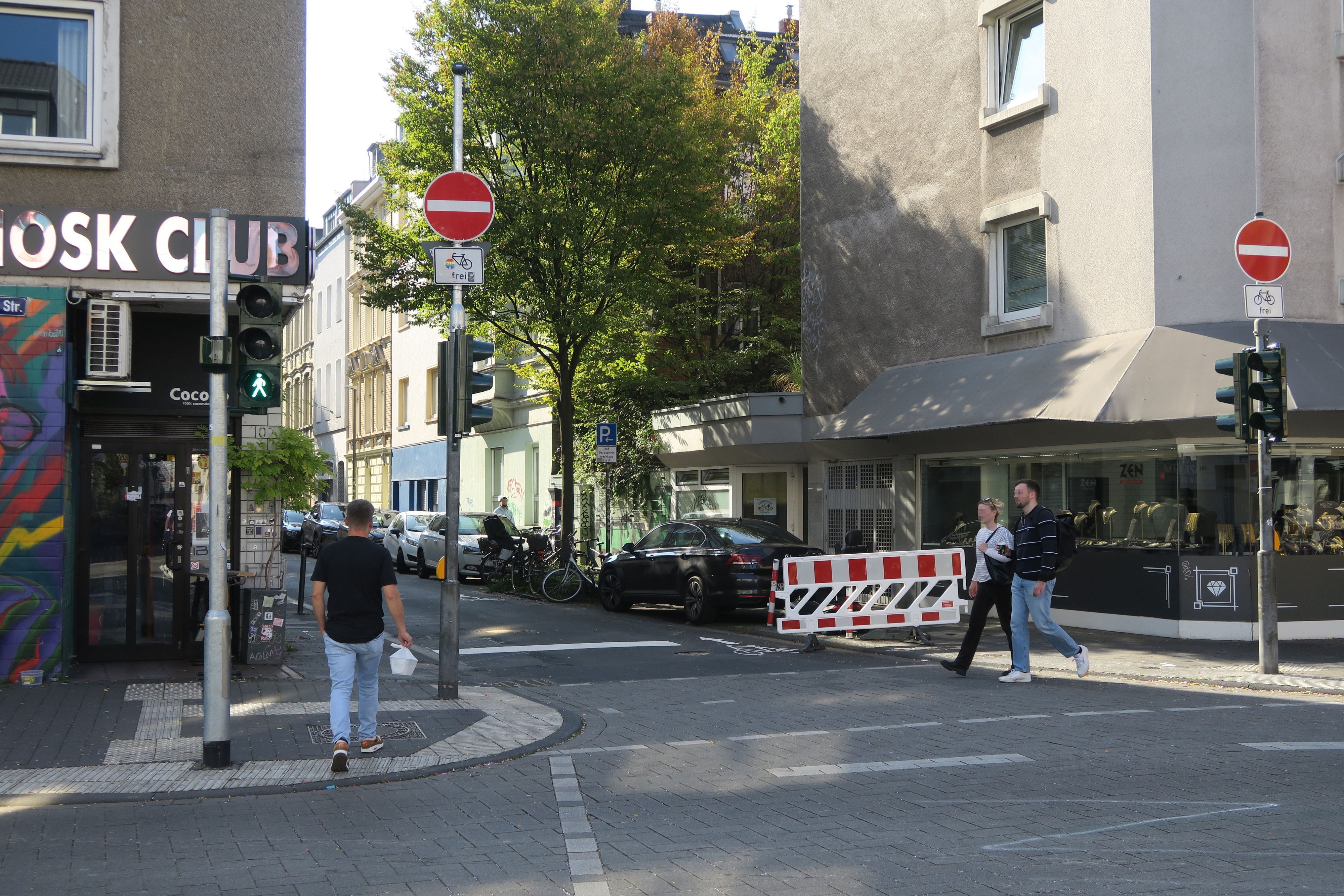 An einer Straßenecke stehen zwei Einfahrt-verboten-Schilder mit Zusatzzeichen „Fahrräder frei“. Im Bild sind außerdem Gebäude mit Geschäften, geparkte Autos, Fußgänger und eine Absperrbake zu sehen.