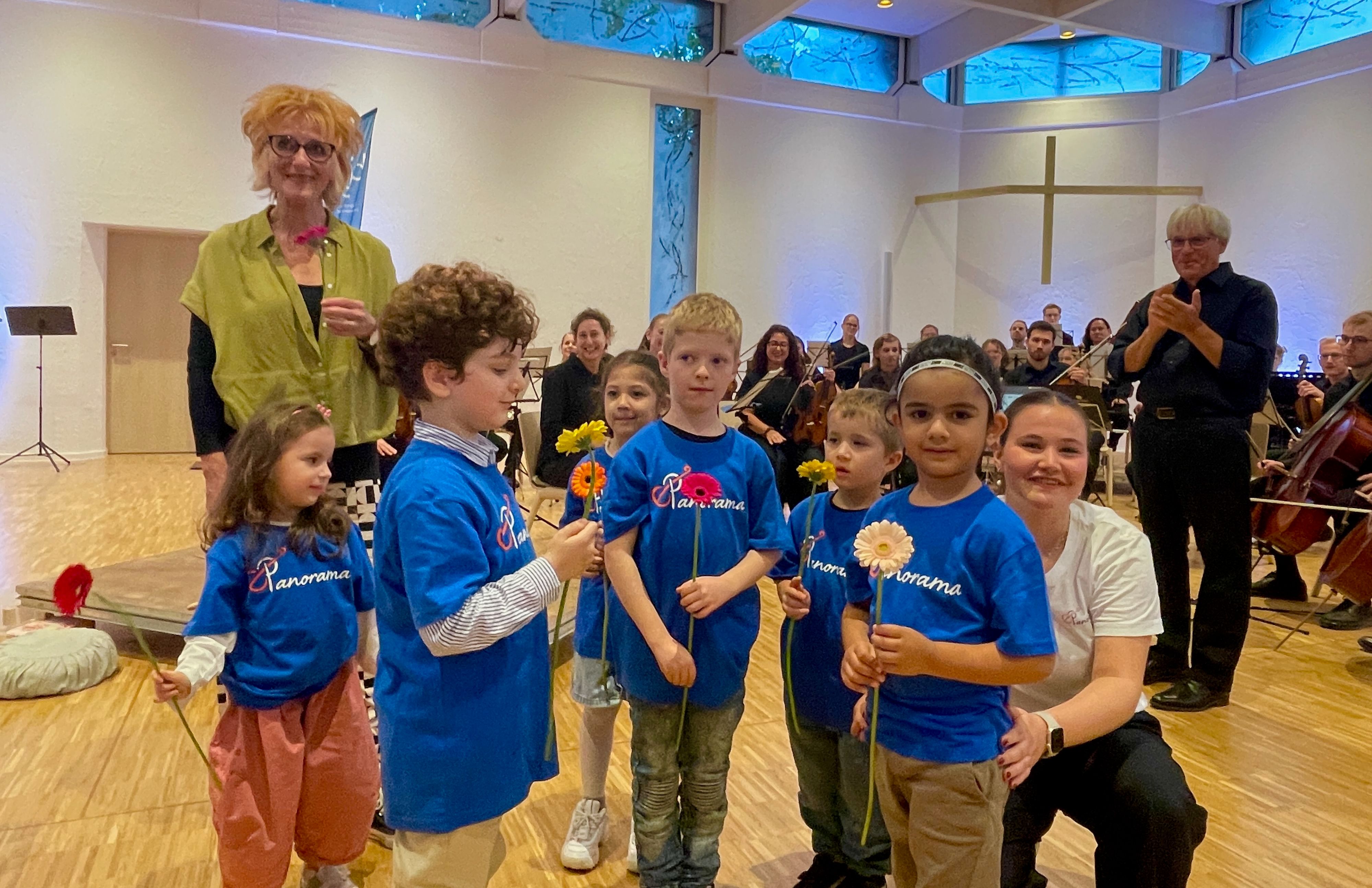 Fünf Grundschulkinder stehen in blauem T-Shirt mit der Aufschrift „Panorama“ und einer Blume in der Hand vor dem CJD-Orchester
