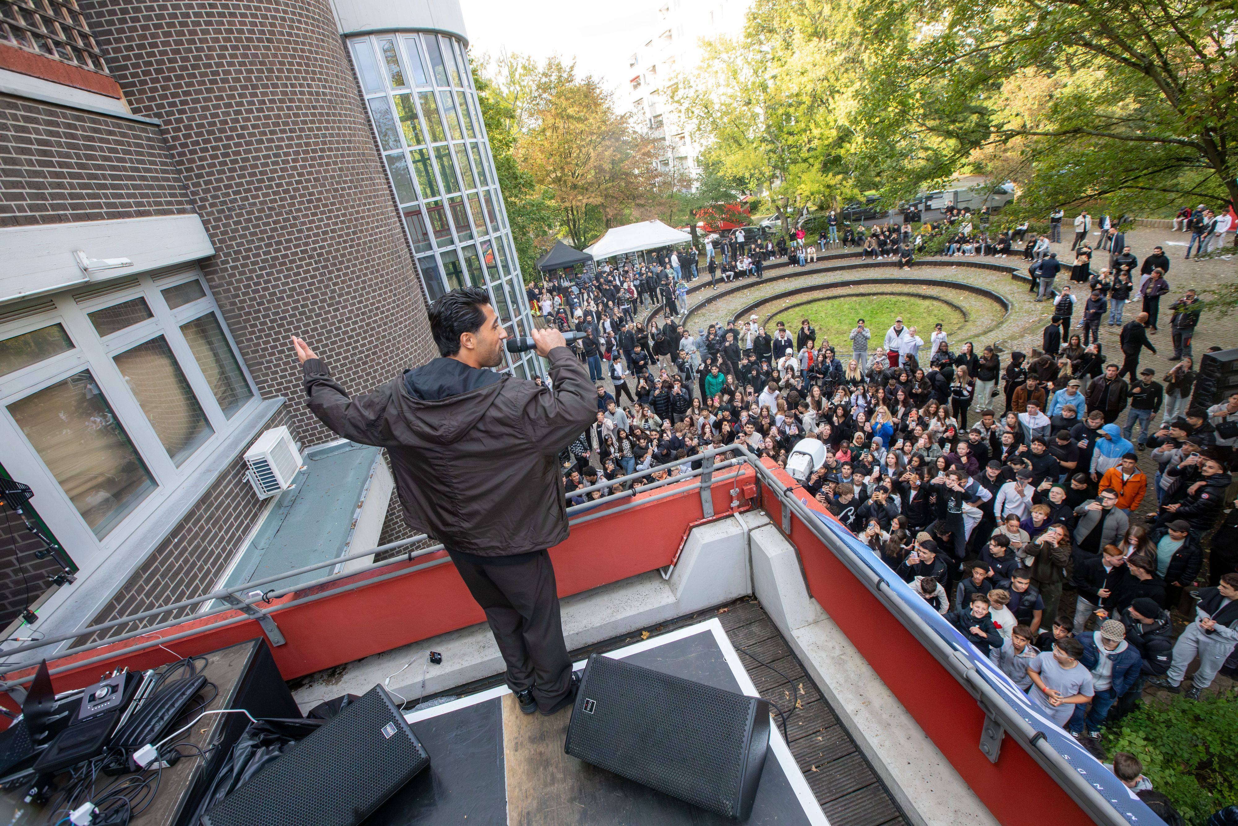 Ein junger Mann singt auf einer Dachterrasse. Unten auf dem Rasen steht eine jubelnde Menge.
