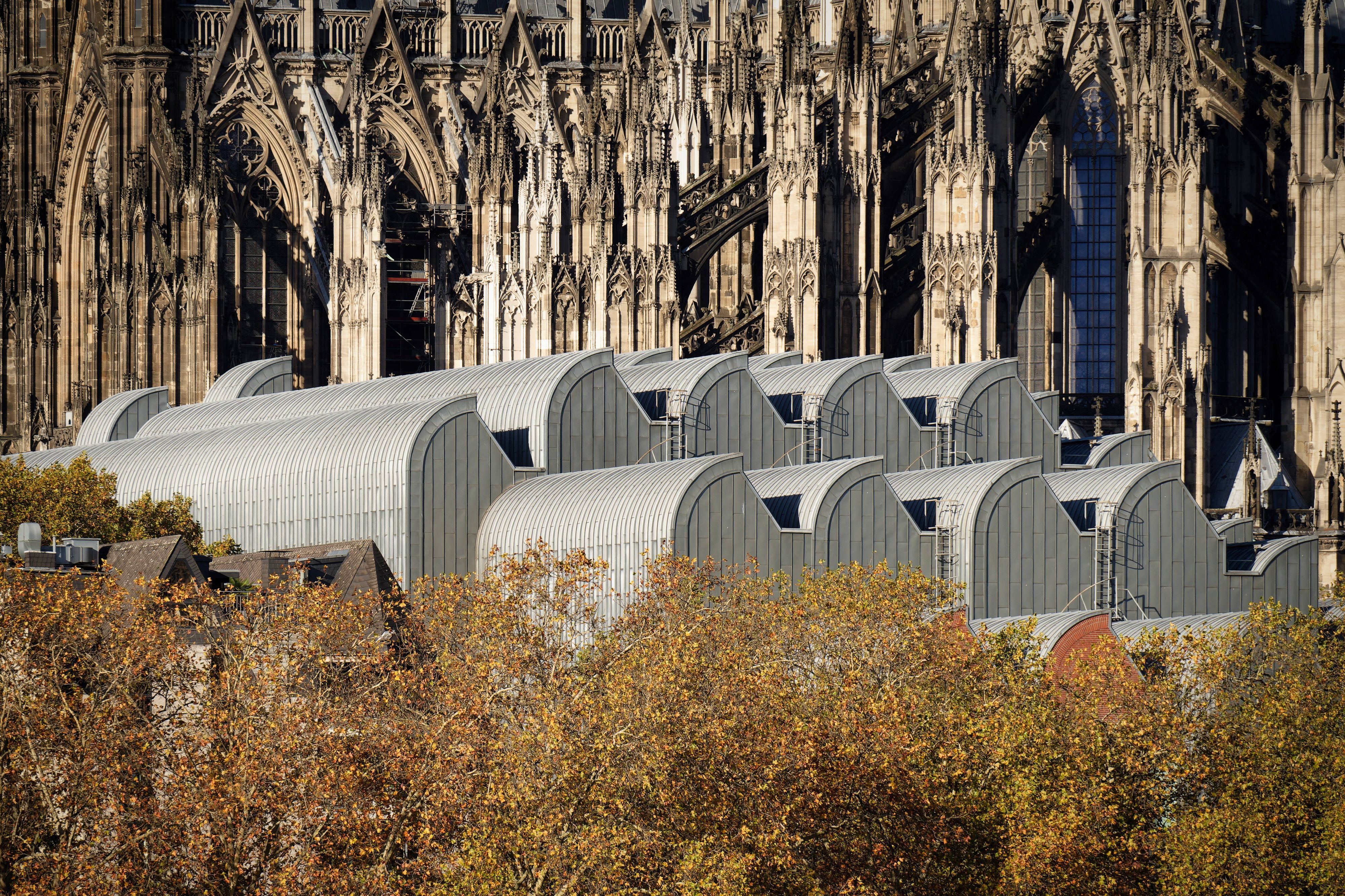 Nahaufnahme der eleganten, wellenförmigen metallischen Dächer des Museum Ludwig, vor der filigranen gotischen Architektur des Kölner Doms und dem herbstlichen Laub.