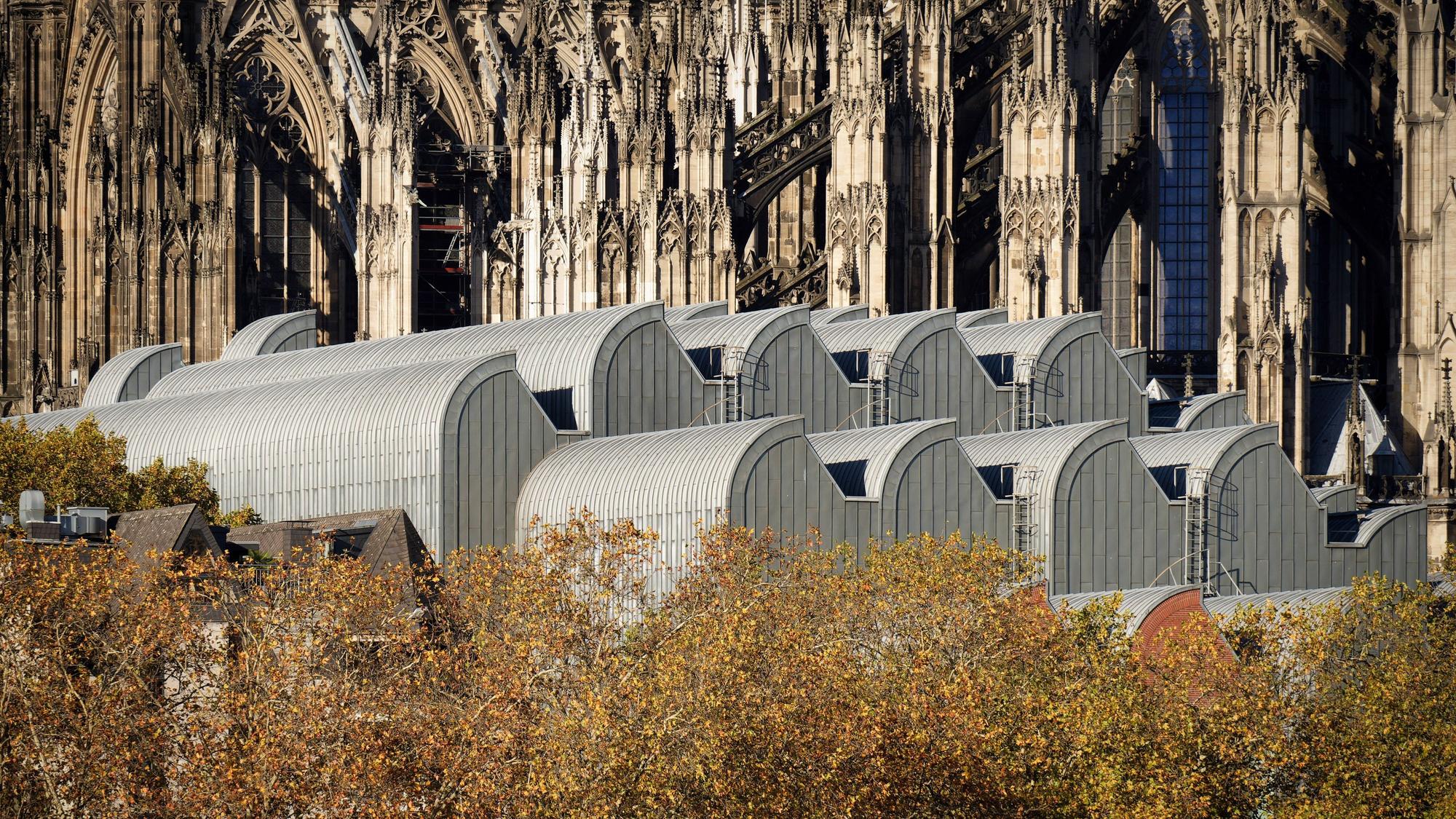 Nahaufnahme der eleganten, wellenförmigen metallischen Dächer des Museum Ludwig, vor der filigranen gotischen Architektur des Kölner Doms und dem herbstlichen Laub.