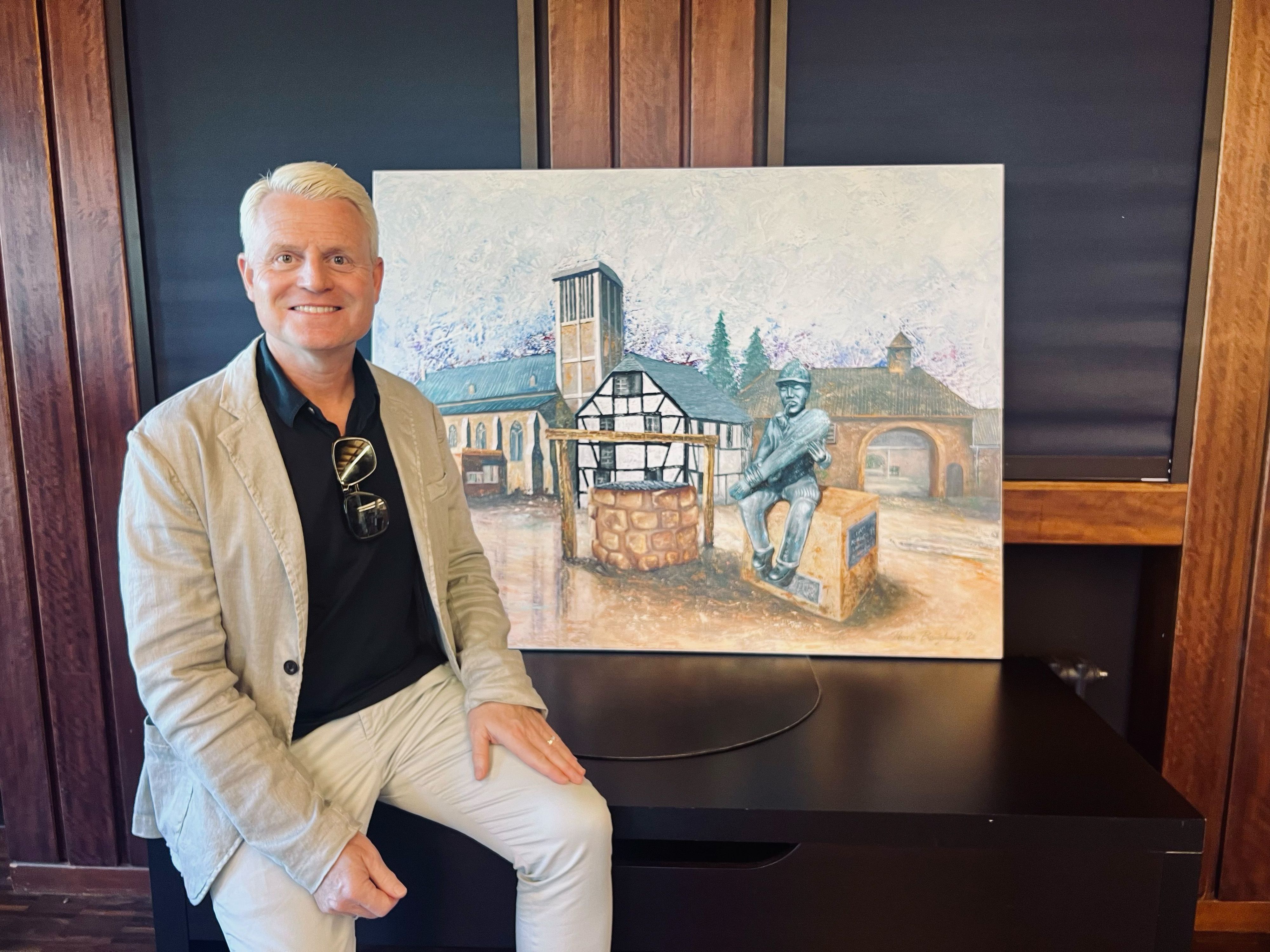 Ein blonder Mann sitzt auf einer Kommode, auf der ein Bild steht vom Besenbinderdenkmal in Eil.