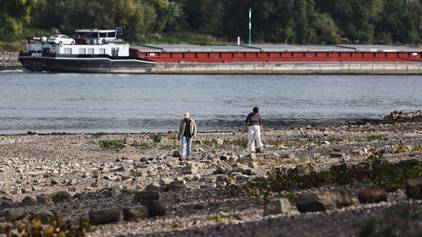 Spaziergänger gehen am Rheinufer in Köln entlang. Im Hintergrund fährt ein Binnenschiff.