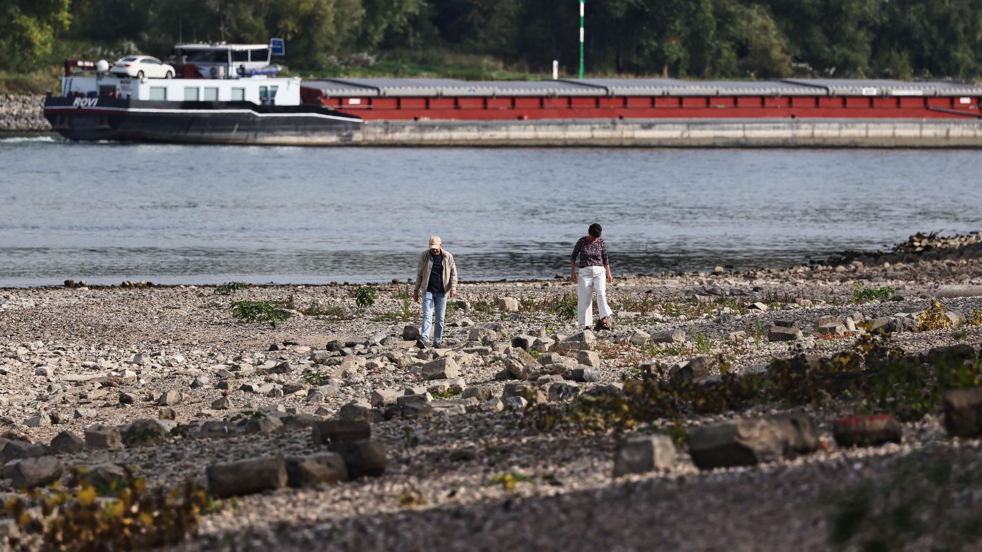 Spaziergänger gehen am Rheinufer in Köln entlang. Im Hintergrund fährt ein Binnenschiff.