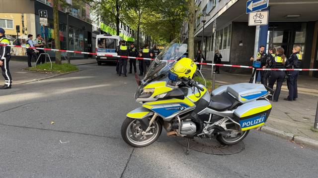 Absperrband hinter einem Polizeimotorrad in der Nähe eines Tatorts. Einsatzkräfte stehen auf einem Parkplatz. Zwei Unbekannte haben mitten in Essen zwei Justizbeamte angegriffen und einen 34 Jahre alten Häftling befreit.