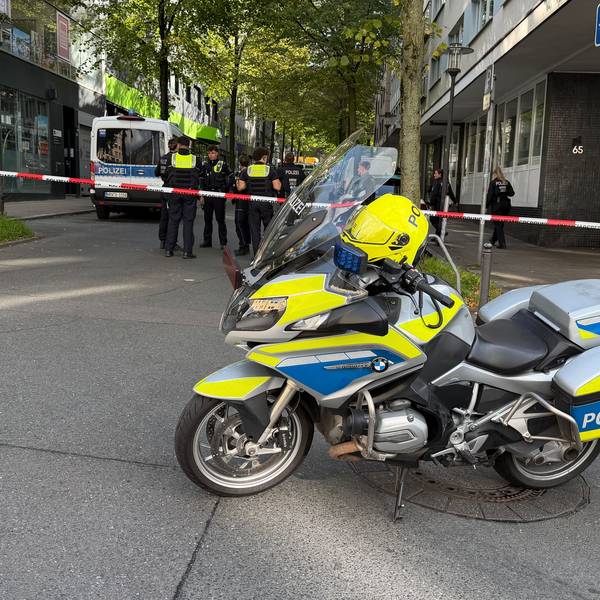 Absperrband hinter einem Polizeimotorrad in der Nähe eines Tatorts. Einsatzkräfte stehen auf einem Parkplatz. Zwei Unbekannte haben mitten in Essen zwei Justizbeamte angegriffen und einen 34 Jahre alten Häftling befreit.